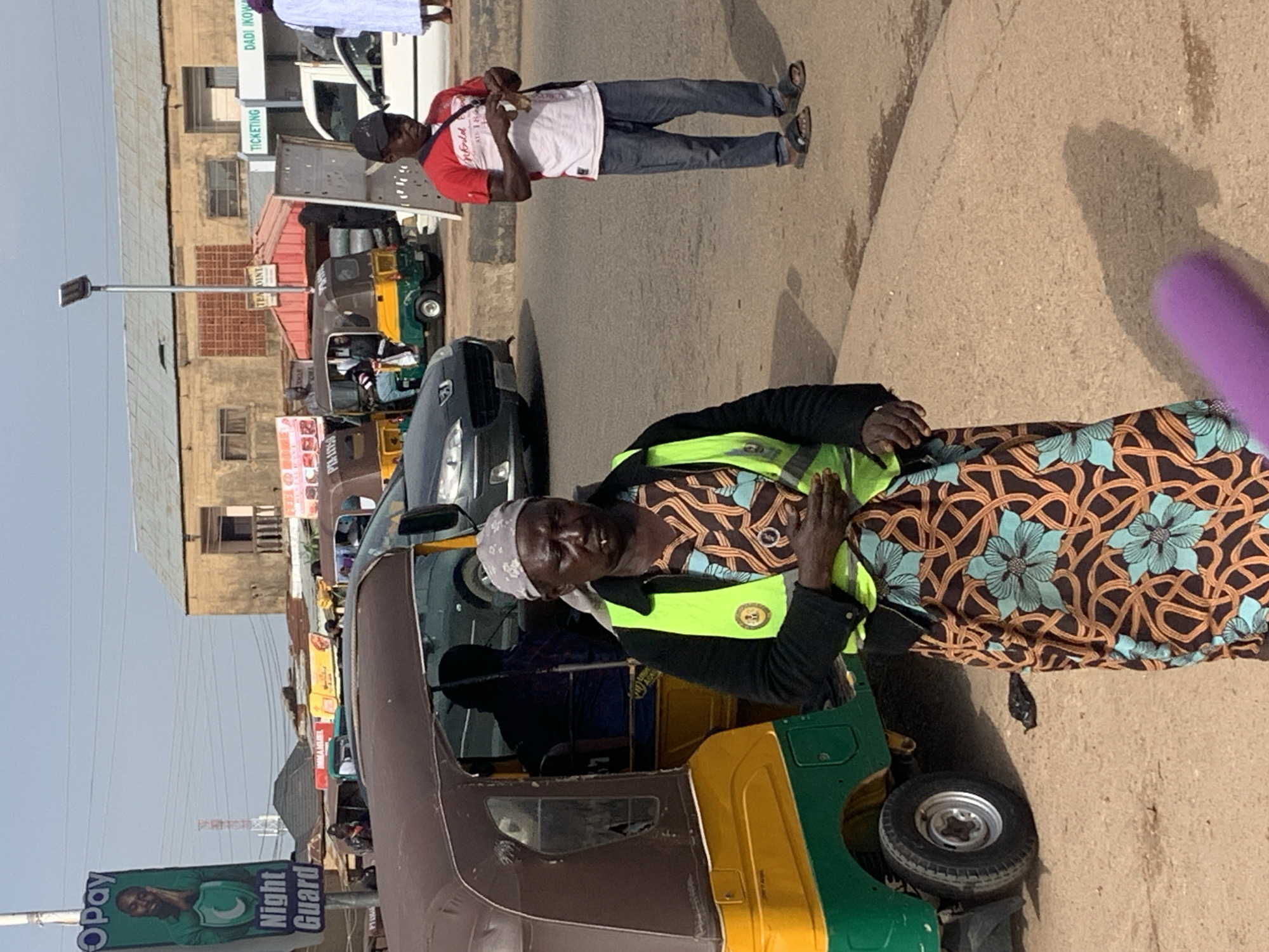 In Gura Top, Nigeria, a woman stands near a colorful auto-rickshaw, her gaze directed downwards. She wears a bright yellow-green vest over a patterned dress and a headscarf. Beside her, a man in a red shirt and blue jeans stands, holding a small object. The backdrop reveals a bustling street with other auto-rickshaws, cars, and buildings, including a billboard for "O Pay Night Guard." The scene captures a slice of daily life in a Nigerian urban environment.