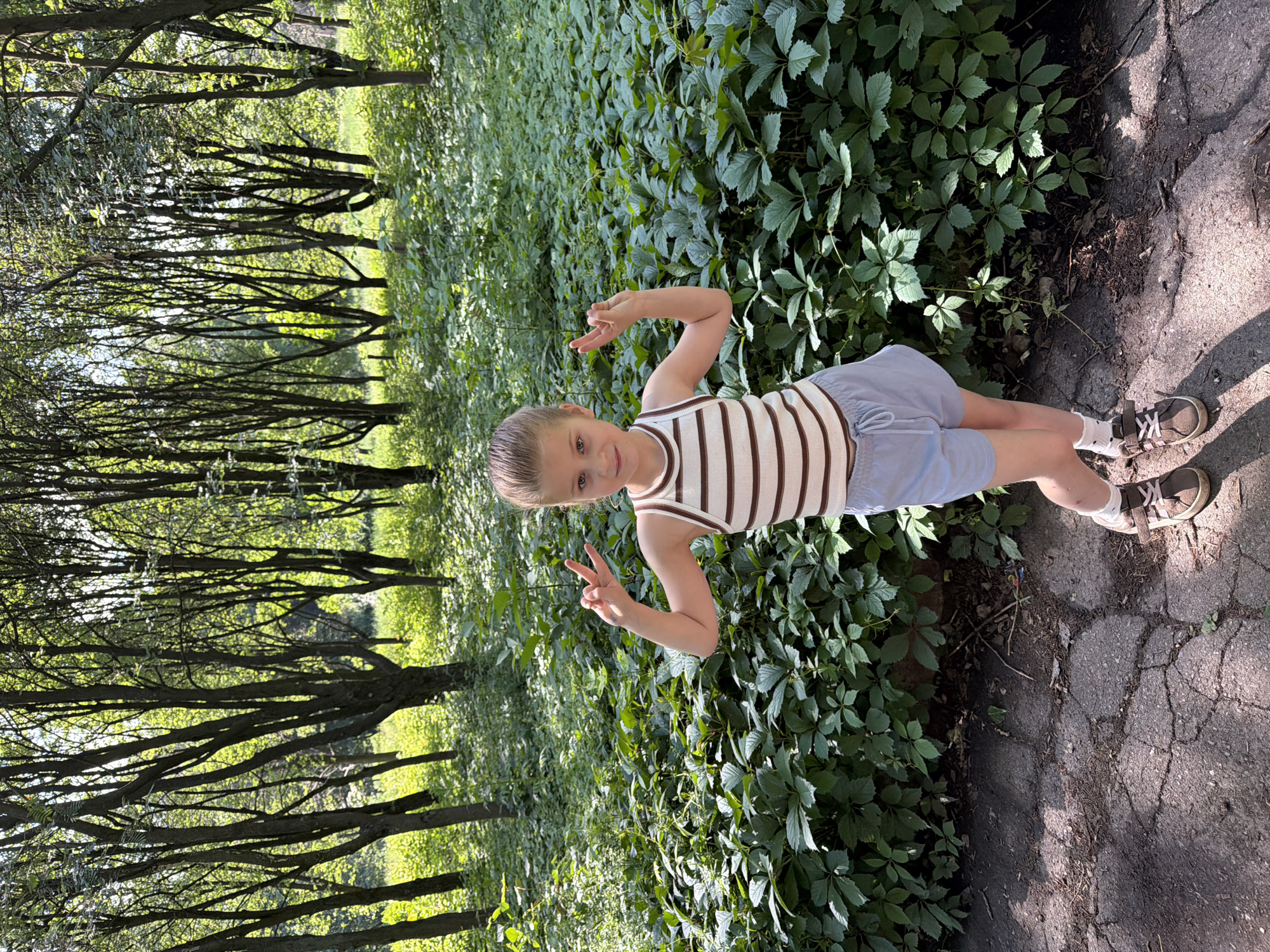 In an outdoor park, a young girl with blonde hair pulled back in a ponytail stands on a cracked asphalt path. She is wearing a striped tank top with brown and white stripes, light blue shorts, white socks, and brown sneakers. The girl is making a peace sign with both hands. She is positioned in front of a dense green shrubbery and a backdrop of trees with sunlight filtering through their leaves.