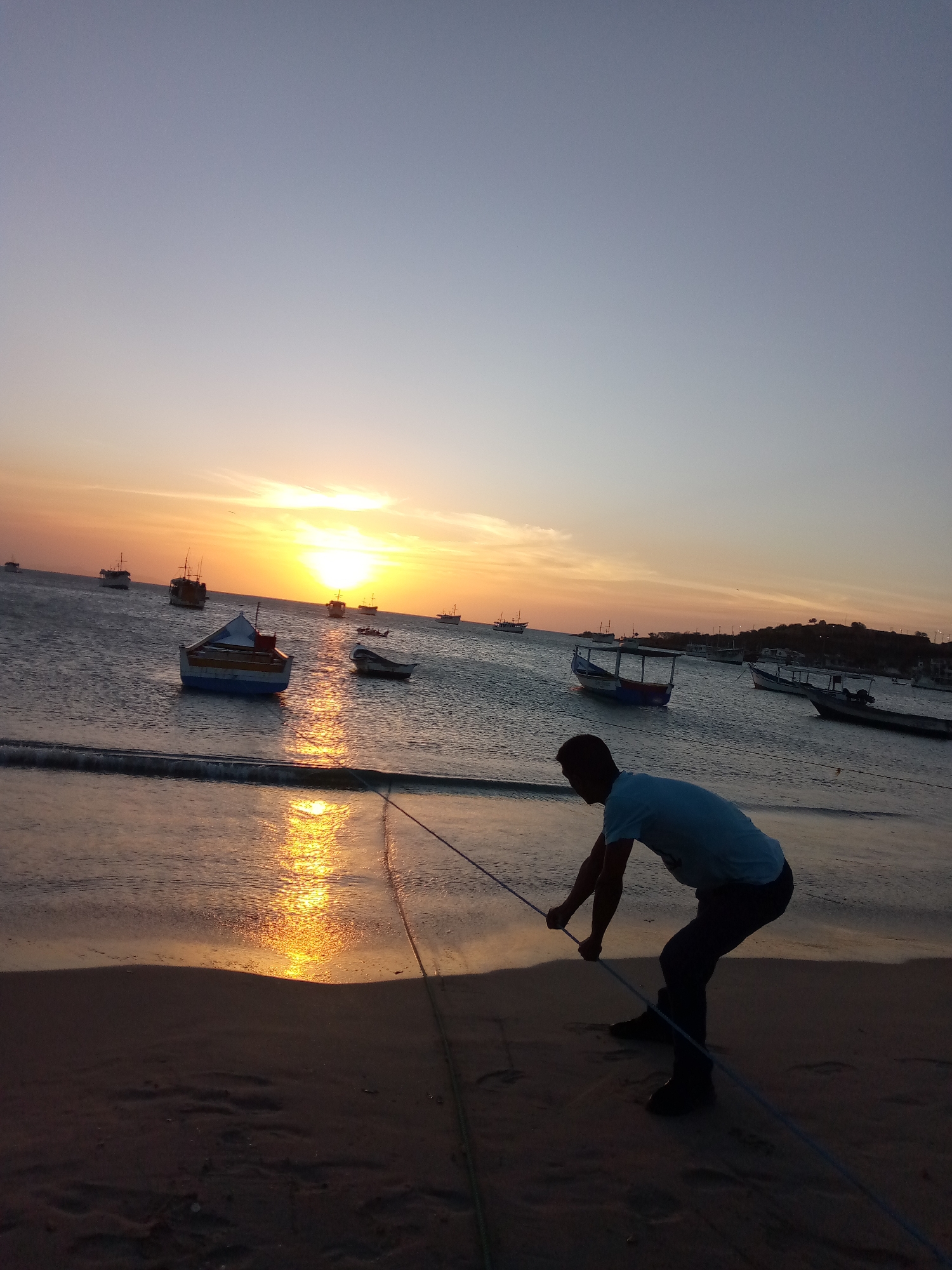 The image shows a person pulling a rope at Playa de Juan Griego, Venezuela, during a sunset. The person is silhouetted, wearing a light-colored shirt and dark pants. Several boats are visible in the water, some close to shore and others further out. The sunset reflects on the water's surface. The sand is a dark color.