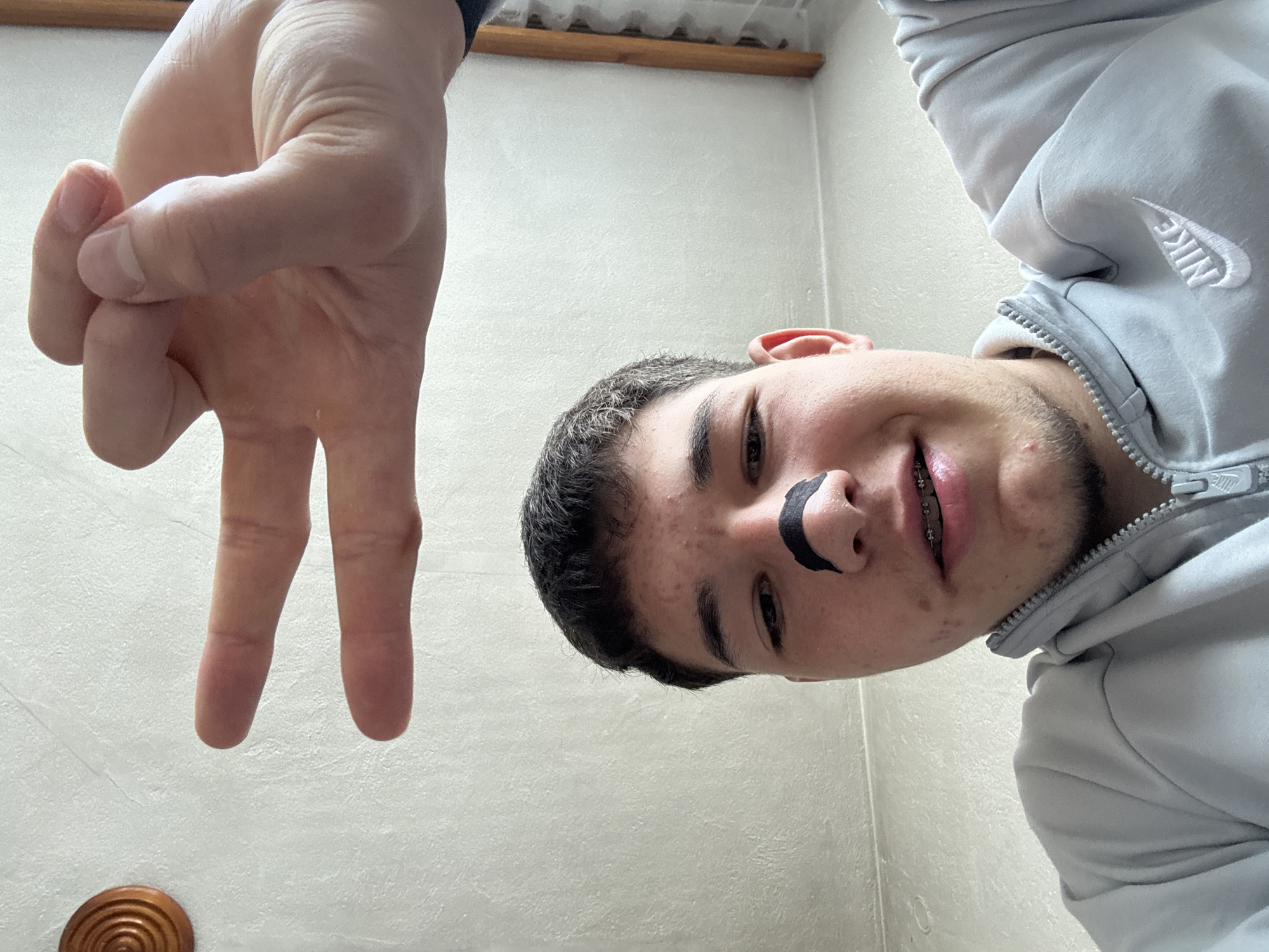 A young man wearing a grey Nike hooded sweatshirt poses for a selfie at eye-level with a white wall as a background. He is smiling with metal braces visible on his teeth. A black crescent-shaped object is located between his nose and upper lip. A hand is making a "V" sign in the upper left corner of the image. In the lower-left corner, there is a small, round, wooden object with concentric circles. The location is unspecified, with no indicators of Zlatograd, Bulgaria.