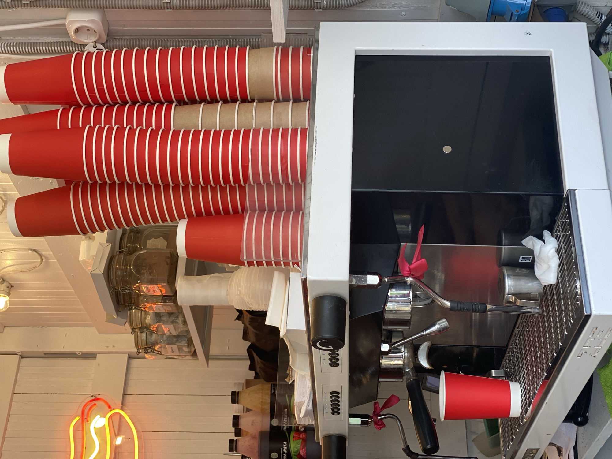The image displays the interior of a beverage and food preparation stall, likely a coffee stand, located in Sochi, Russia. The environment features vertical white wooden paneling walls.
In the foreground and midground, a professional-grade espresso machine with a white top, black front panel, and chrome components dominates the scene. Two portafilters are attached, and two steam wands, adorned with small red ribbons, extend from the machine. A single red paper cup with a white rim rests on the machine's metal drip tray, which also displays the partial brand name "SOS" or similar. A small metal pitcher and a crumpled white paper napkin are also visible on the drip tray.
Above the espresso machine, multiple tall stacks of red paper cups with white interiors and rims are arranged on a white shelf. Some stacks incorporate plain brown paper cups. Further back on shelves, several clear glass jars with metal clamp lids contain various dry ingredients. To the left, several plastic bottles of liquid syrups or sauces are present, with one black-labeled bottle clearly showing "МАЛИНА-МЯТА" (Raspberry-Mint) and another visible label reading "ИМБИРНЫЙ" (Ginger). Behind these bottles, dark bags, possibly containing coffee beans, are stacked. Clear plastic cups or lids are also organized on the shelves.
On the left wall, a yellow-orange neon sign shaped like a hot dog with mustard is illuminated. The upper right portion of the image reveals a white electrical outlet and grey corrugated electrical conduit running along the wall. A bare light bulb socket with a bulb is visible on the upper left wall. No people or active interactions are depicted in the scene, which appears functional and prepared for service.