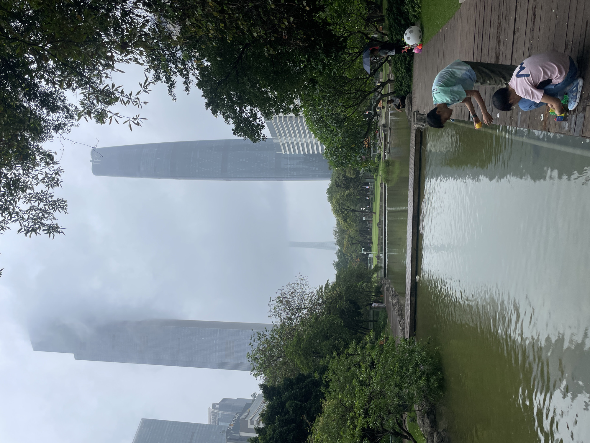 The image depicts an outdoor scene in Tianhe District, China, characterized by a murky atmosphere and modern architecture.
Two children are kneeling on a wooden platform beside a body of water. One child wears a light green and blue shirt, while the other wears a pink shirt with a number "7" on it. They appear to be playing near the water's edge.
The backdrop features a row of trees along the water’s edge and several prominent skyscrapers shrouded in fog. A modern building with white stripes can be partially seen among the trees. The Canton Tower is visible but obscured by fog. Additional buildings are also present, including one that appears to say "GRAND PLAZA" on its side.
A bicycle, possibly belonging to an adult, is parked on the wooden platform, near the children. The overall impression is that the weather is overcast or foggy, reducing visibility of the city skyline.