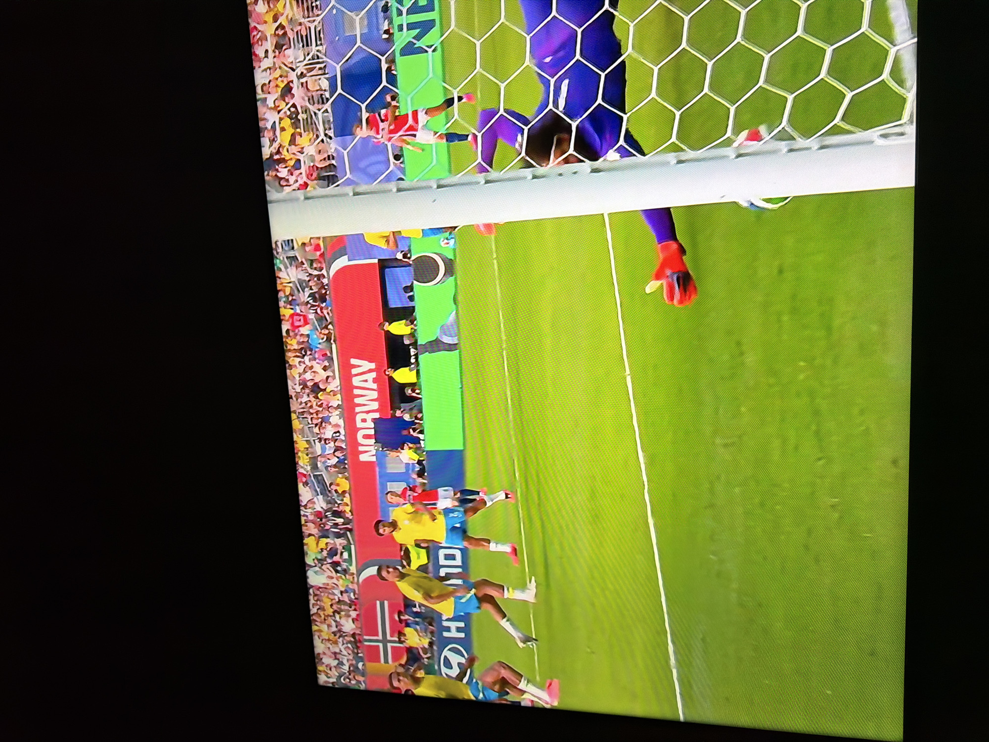 The image shows a soccer game on a bright green field. In the foreground, a goalkeeper in a purple uniform is diving to save a goal. A soccer ball is visible partially in the goal net. In the background, players in yellow and red uniforms are on the field, and spectators are in the stands. A banner with "NORWAY" is visible in the background. The scene is captured from a slight upward angle, likely from a spectator's perspective. The location in Ognianovo, Bulgaria is not depicted in the image.