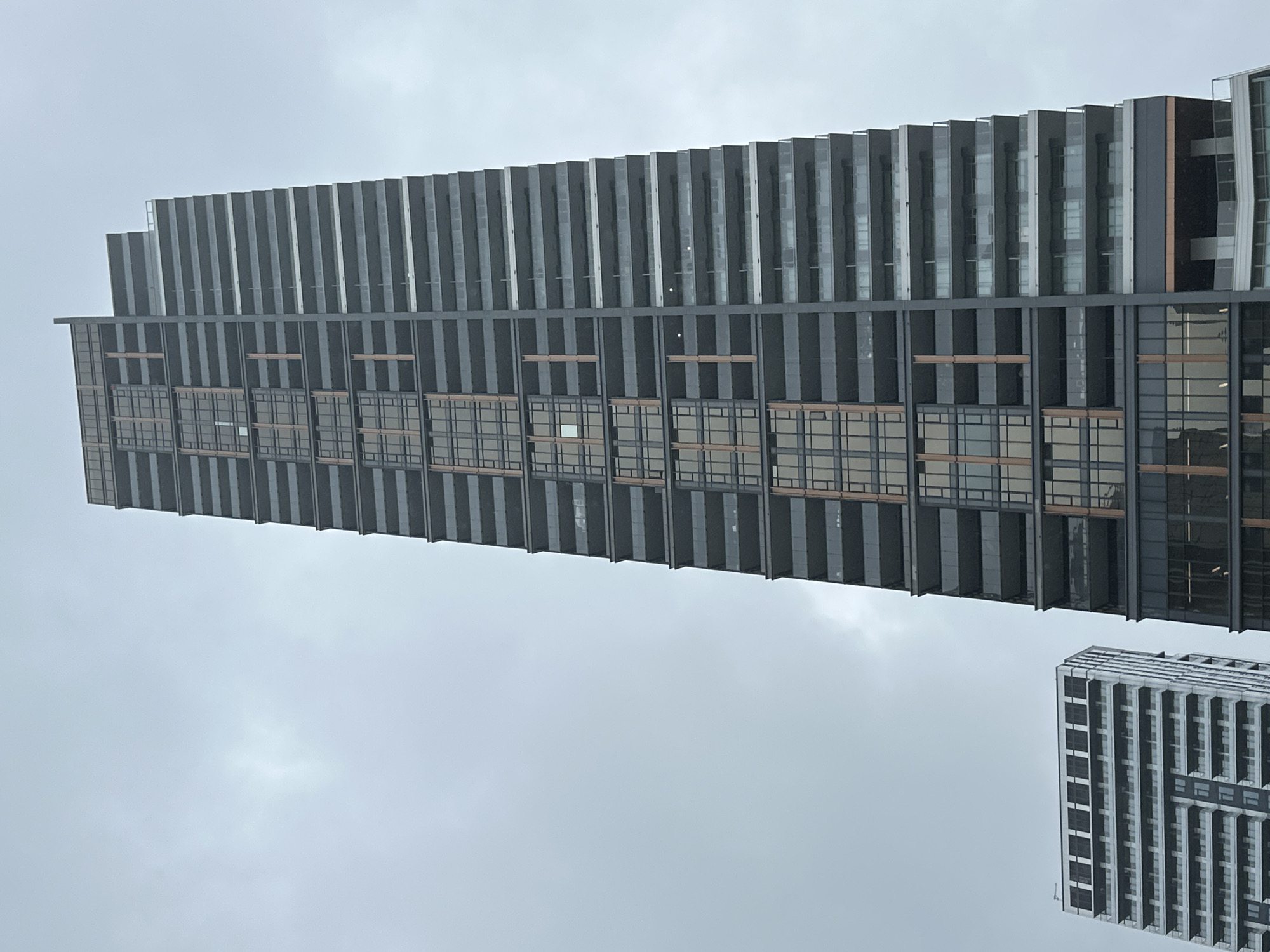 The image shows two modern apartment buildings in Burnaby, Canada, against a cloudy sky. The building on the right is a tall, dark-colored skyscraper with a facade featuring alternating sections of dark grey panels, glass windows, and thin, vertical bronze-colored accents. Multiple balconies with glass railings are visible on this building. The building on the left is shorter and lighter in color, with a repeating pattern of white balconies and dark-framed windows. Both buildings appear to be residential structures. The overall lighting suggests an overcast day.