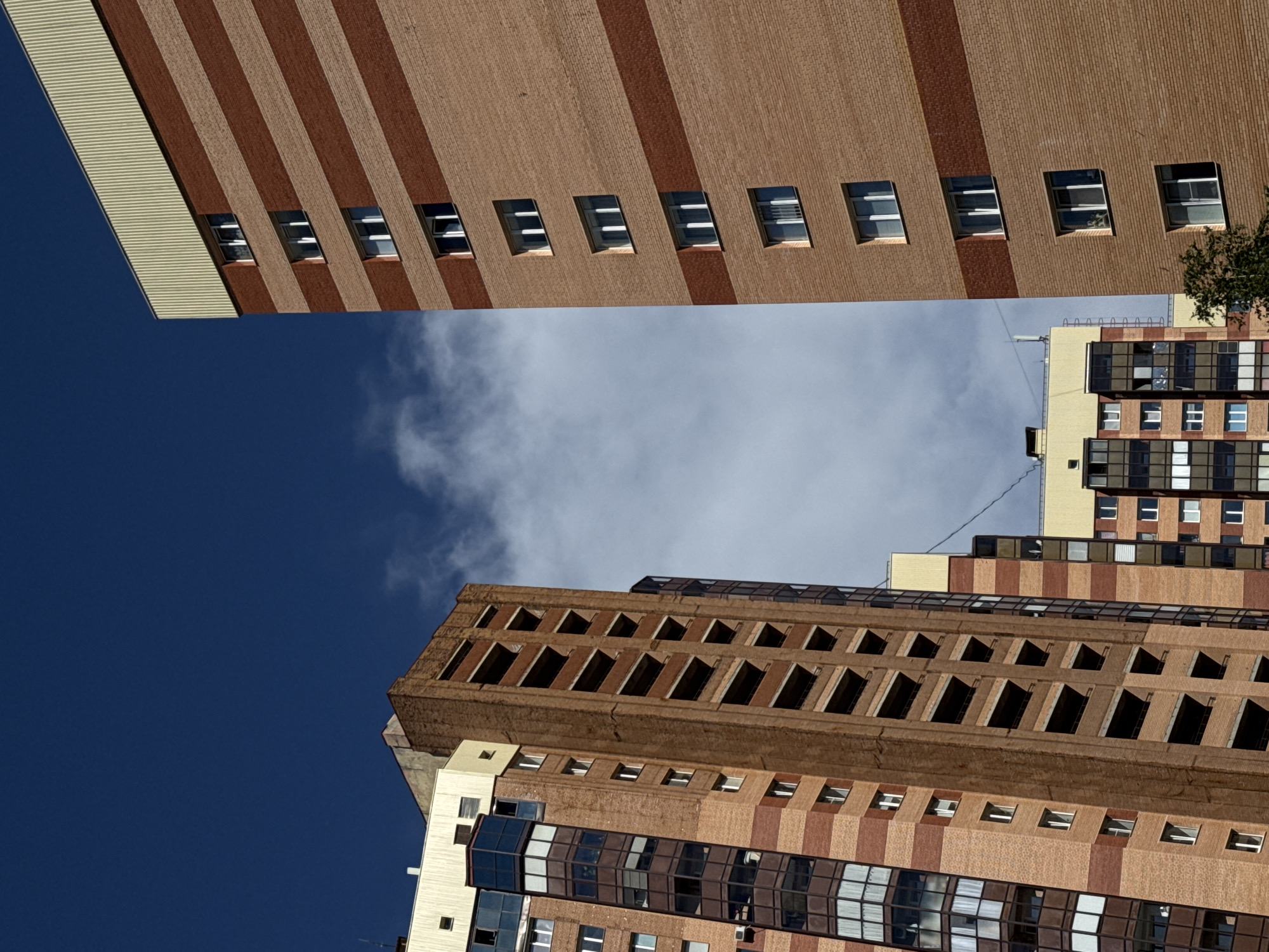 The image displays an urban scene featuring multiple residential high-rise buildings against a clear, deep blue sky with scattered white clouds. The perspective is a low-angle shot, emphasizing the height of the structures.
On the left, a multi-story building with a light brown brick facade is visible, featuring alternating bands of reddish-brown brick and numerous glass-enclosed bay windows or balconies that extend vertically. The upper section of this building incorporates cream-colored panels. To its right and slightly behind, a taller, narrower building constructed of darker brown brick appears to be either unfinished or abandoned, characterized by a series of open, recessed rectangular voids stacked vertically without windows or railings.
Dominating the right side of the frame is another tall, multi-story building with a light tan brick facade. This building is distinguished by horizontal bands of reddish-brown brick at regular intervals, approximately every two to three floors. It features numerous rectangular windows with white frames. The roofline is capped by a distinct light beige or yellow corrugated metal paneling.
In the lower middle-right, partially obscured by the foreground buildings, are elements of additional residential structures with similar brick facades and window patterns, some topped with yellow or cream-colored sections. A small cluster of green foliage is visible at the very bottom right corner. The sky is predominantly deep blue with patches of white cumulus clouds. No people or discernible actions are present in the scene. The location is the city of Unknown, Unknown.