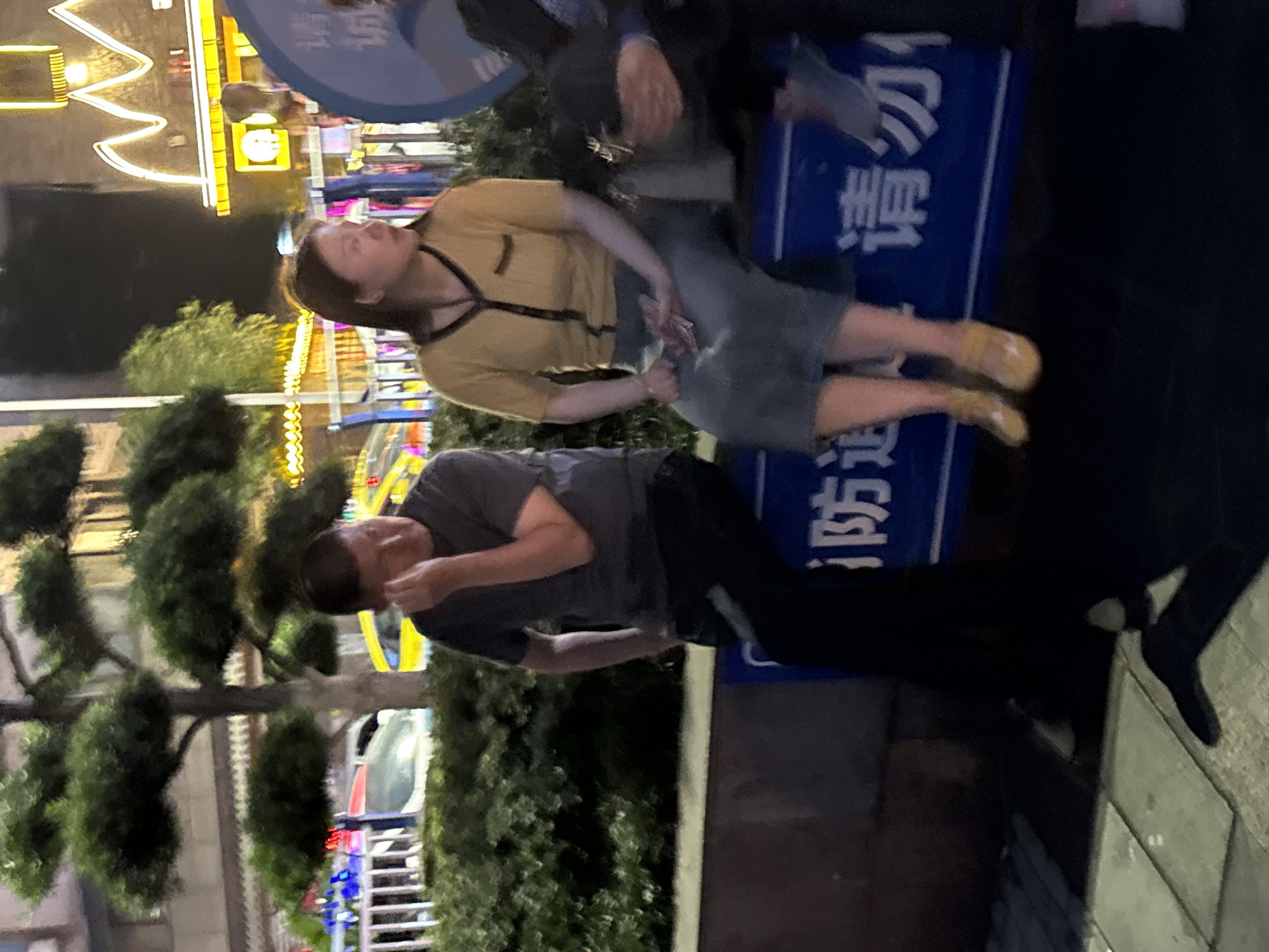 The image shows an outdoor scene, likely at night in the Yuzhong District of China, given the Chinese characters on a blue sign.
A man in a gray t-shirt and black pants stands on the sidewalk, his hand near his face, possibly on a phone.
A woman in a yellow top and gray skirt is standing next to a dark colored barrier that has white letters on a blue background.
A person is near the barrier, facing forward.
There are trees, building lights, and railings in the background, which suggests a bustling urban environment. The lighting is bright, indicating street or building illumination.