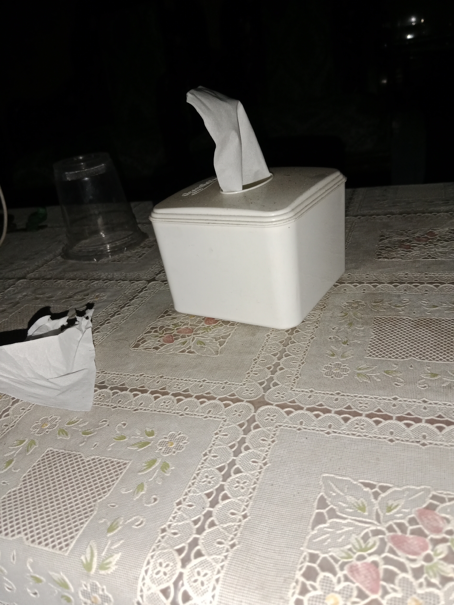 The image shows a close-up view of a table covered with a white lace tablecloth. On the table, there is a white tissue box with a tissue pulled out. To the left of the tissue box, there is a clear plastic cup, upside down. In the foreground, on the left side, there is a crumpled white tissue. The background is dark and out of focus, suggesting it is nighttime and the setting is indoors or outdoors with minimal artificial lighting. The scene is devoid of any individuals or any discernible activity. The image does not provide any specific information that would indicate its location in Ringinrejo, Indonesia.