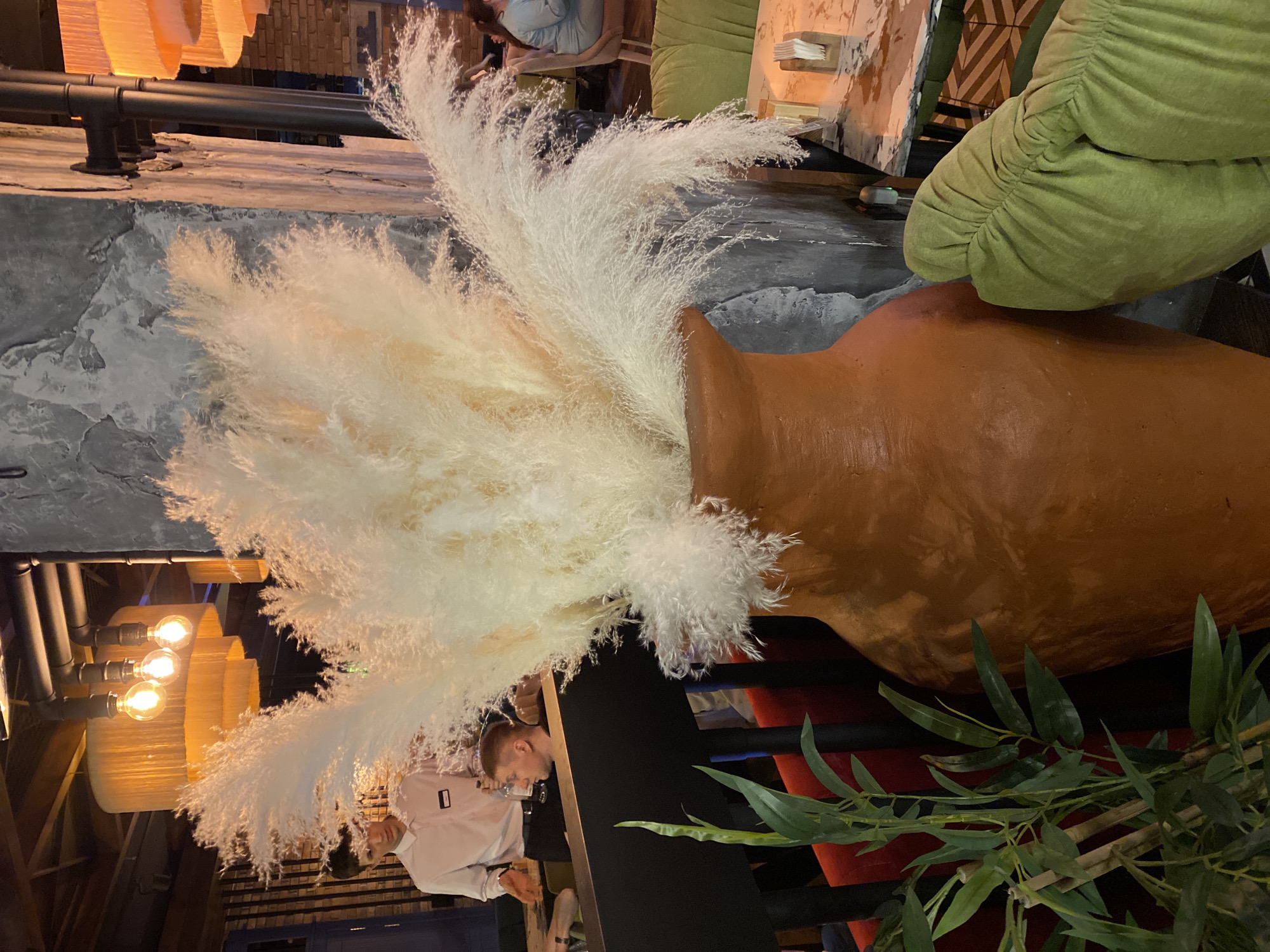 The image depicts an interior scene, likely in a restaurant or cafe. A large, tan-colored clay pot is the focal point, filled with tall, white, feathery pampas grass. To the right of the pot, a green, cushioned chair or sofa is visible. In the background, there are other tables and chairs, suggesting a dining area. A person wearing a white shirt is partially visible near the lower left of the frame. Above them is a light fixture with multiple bulbs encased in woven shades. To the right of the person is foliage with green leaves. The scene is likely in a modern, stylish establishment, as indicated by the decor and lighting. The location information (Yekaterinburg, Russia) provides context for the cultural and architectural style of the space.