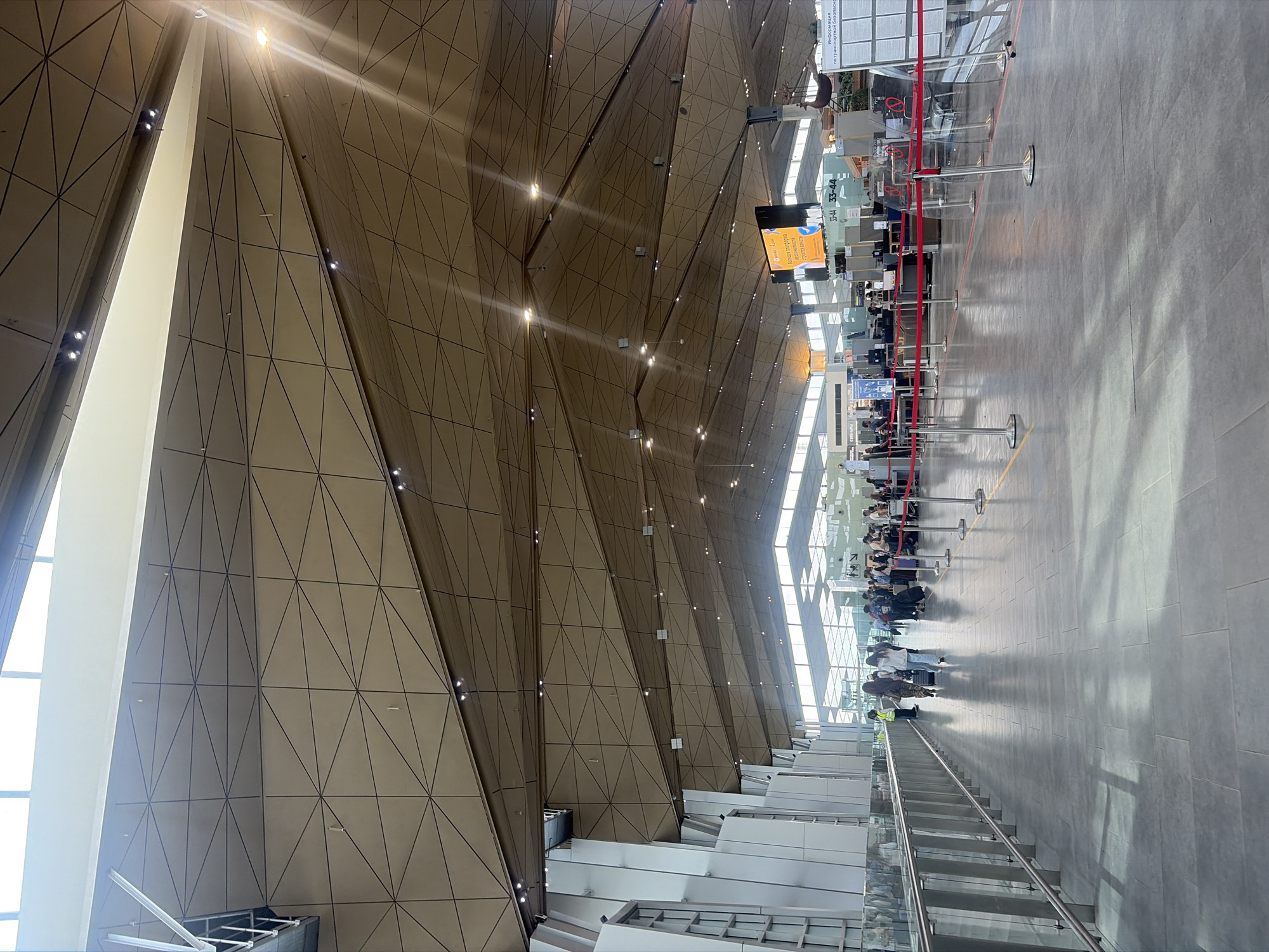 The image captures the interior of a spacious, modern airport terminal with a distinctive architectural design. The most prominent feature is the dramatic, faceted ceiling, composed of numerous large, geometric panels in light brown or beige tones, accented by dark lines forming triangular and quadrilateral patterns. Recessed spotlights are embedded within these panels, casting bright beams and reflections. Large windows or skylights are also integrated into the upper structure, providing ample natural light.
Below this expansive ceiling, a wide, light-colored tiled floor extends into the distance. A queue of passengers is visible in the midground, standing behind red stanchion ropes that delineate the waiting area. These individuals, mostly standing and some accompanied by luggage, appear to be waiting for check-in or security procedures. Several check-in counters or service points are discernible further along the queue, equipped with digital screens and airport machinery. An airport staff member in a high-visibility yellow vest is also present near the queuing area. To the left, a modern staircase with glass railings ascends, suggesting access to different levels within the terminal.
The scene depicts a bustling, yet orderly, daytime airport environment. Several signs with visible text are present. A prominent digital display screen shows Cyrillic script, including "ПРОГРАММА ПОДДЕРЖКИ" (SUPPORT PROGRAM). Other signs display numbers such as "33-44," likely indicating gate or counter numbers, and text related to "Информация по транспортной безопасности" (Information on transport security). The terminal exhibits a clean, well-maintained, and architecturally striking appearance, indicative of a major modern transportation hub.