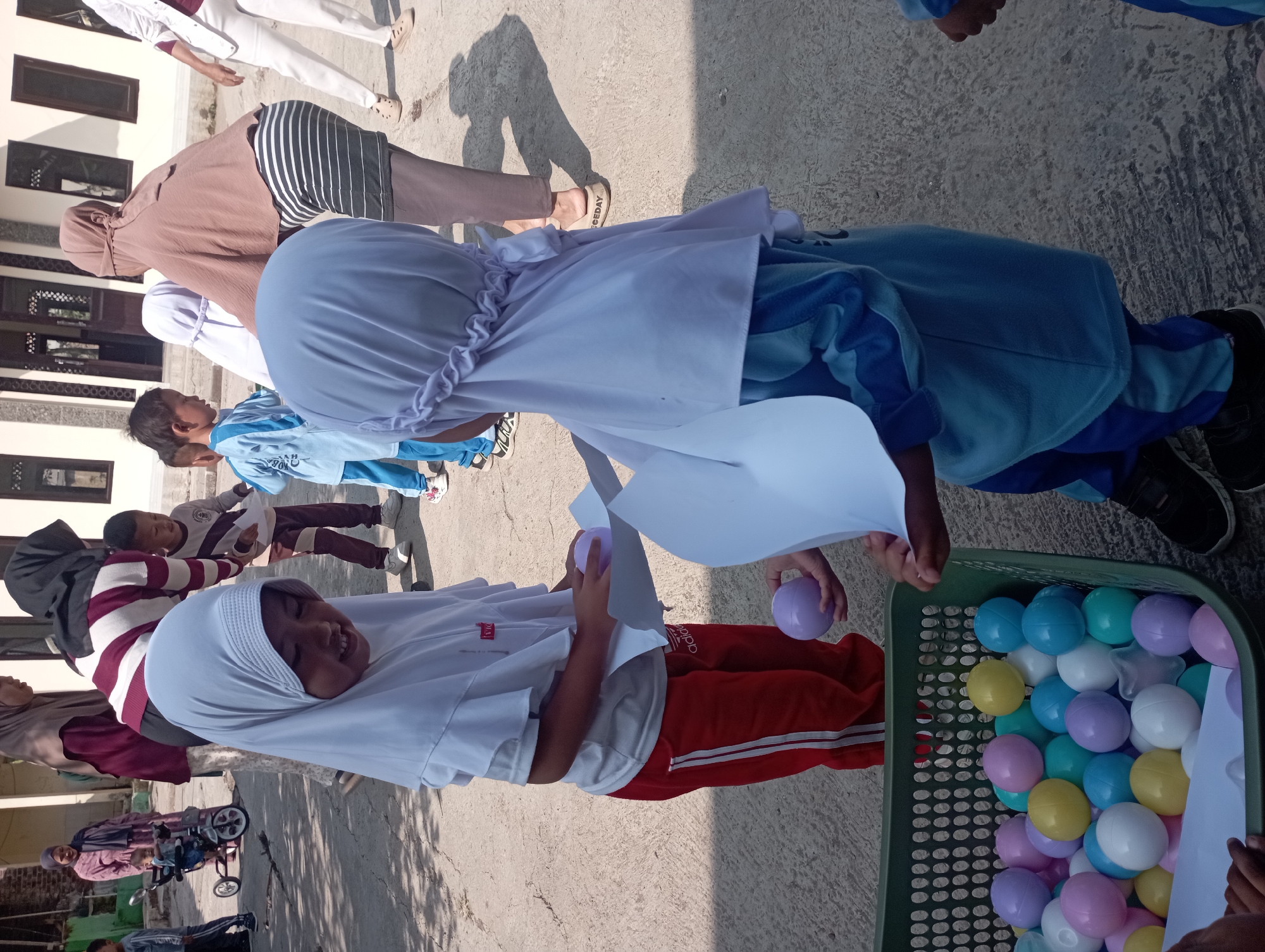 In Garut, Indonesia, a group of children are participating in an outdoor activity, likely a game involving a basket of colorful plastic balls. In the foreground, two young girls wearing hijabs are interacting. The girl on the left, in a white hijab and red pants, is holding a piece of white paper and a light purple ball. The girl on the right, in a light blue hijab and uniform, is also holding a piece of white paper and appears to be receiving something from the girl on the left. Behind them, other children and adults are visible. One adult, wearing a brown hijab and a striped skirt, is standing and looking down. Another child, wearing a blue and white shirt, is standing nearby. In the background, other individuals are seen, including a woman pushing a child in a stroller. The environment appears to be an outdoor paved area, possibly a schoolyard or a community space, with a building visible in the background. The overall scene depicts a playful and communal atmosphere.