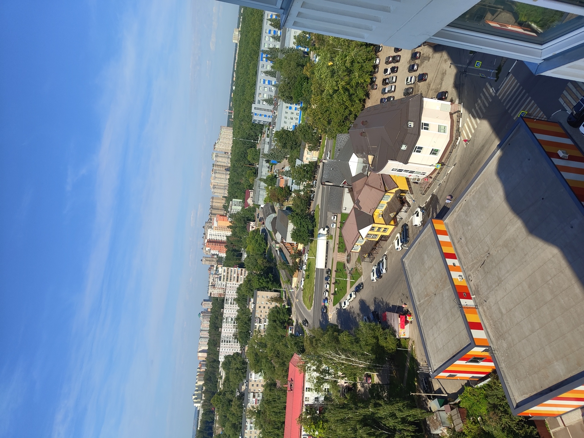 The image shows an aerial view of a residential area in Ramenskoye, Russia, on a sunny day. In the foreground, two modern buildings with flat roofs are visible. The building on the left has a facade with vertical stripes of orange, yellow, red, and white. The building on the right is partially visible and has a dark roof and light-colored walls.
A street runs through the scene, with several cars parked along its sides and a crosswalk. Several smaller buildings, including houses with yellow and brown facades, are situated along the street. Further in the background, a large, multi-story building with blue accents and numerous windows is visible. Beyond that, a dense green forest stretches across the landscape, followed by a line of tall, apartment buildings and in the far distance, more high-rise buildings. The sky is a clear blue with a few wispy clouds.
In the middle ground, a white truck is driving on the road. A few people are walking on the sidewalks and near the buildings. The overall scene depicts a typical urban environment with a mix of residential buildings, commercial structures, and natural greenery.