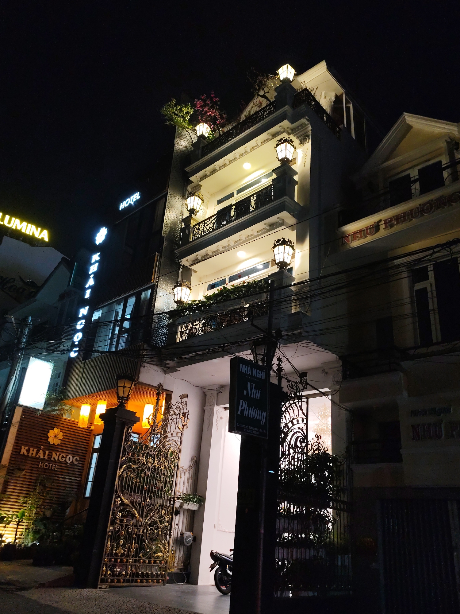 A nighttime shot captures two adjacent buildings, likely hotels or guesthouses, in Da Lat, Vietnam. The building on the left, marked "KHAI NGOC HOTEL" in neon lights and also in a lit sign with a yellow flower, has a modern, vertical sign and bright lights. A black metal gate adorned with intricate designs stands at the entrance. The building to the right, "NHU PHUONG," is a few stories high and features lights on balconies. A sign is visible near its entrance. Power lines are strung across the scene. A motorcycle is parked near the entrance of "NHU PHUONG."