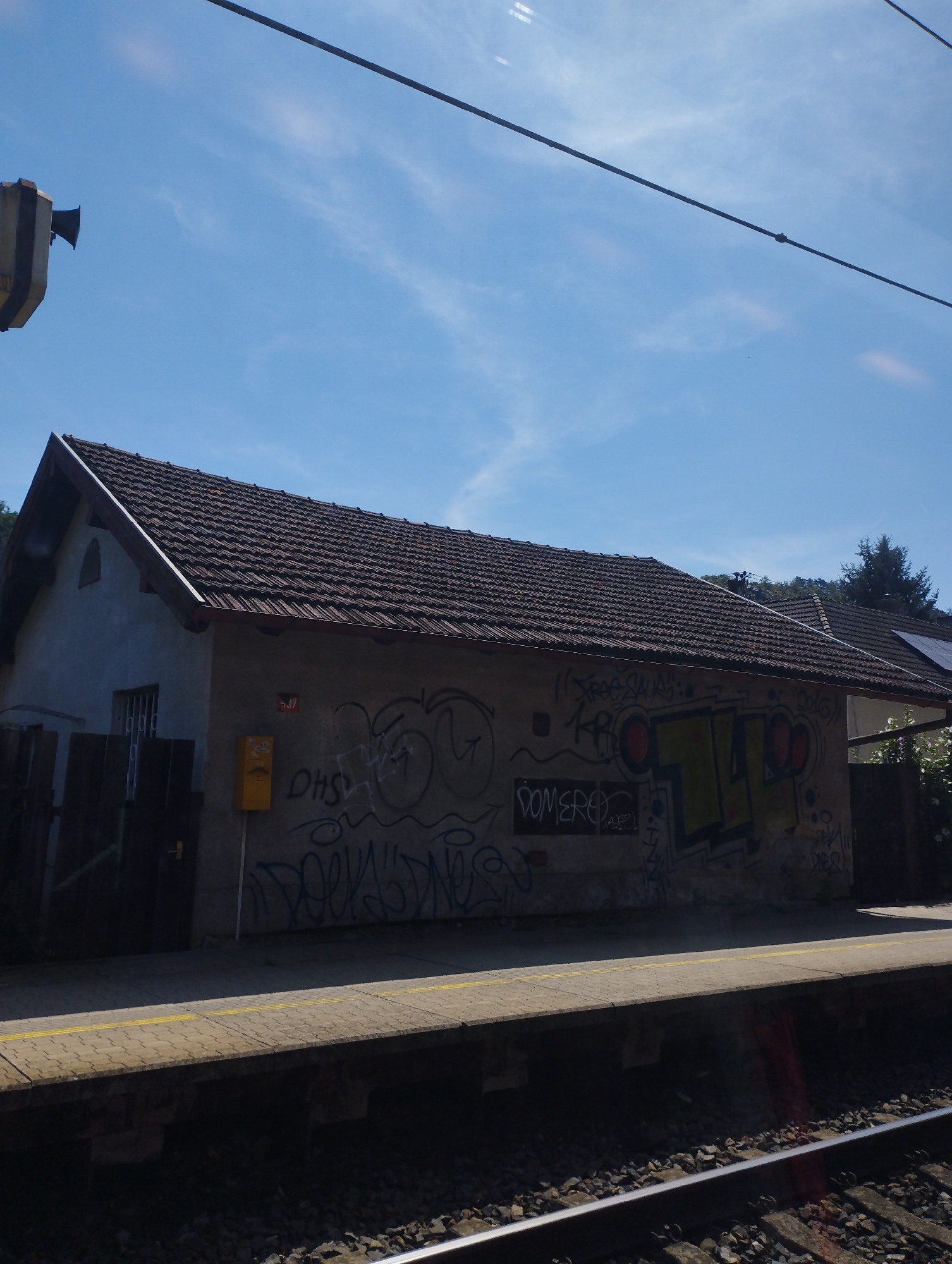 The image shows a low-rise building with a tiled roof, covered in graffiti, on a sunny day with a blue sky and scattered clouds. The building appears to be a small train station or an outbuilding next to a railway line, as indicated by the presence of a platform with yellow lines and train tracks in the foreground. To the left of the building, there is a metal fence and a window with bars. A yellow mailbox is mounted on the wall of the building. The building's wall is heavily graffitied with various tags and designs, some in colorful spray paint. Behind the building, trees and foliage are visible, suggesting a somewhat rural or suburban setting. A wire runs diagonally across the upper portion of the image, possibly an overhead power line for the train. The overall impression is one of urban decay or neglect due to the graffiti and the weathered appearance of the building.