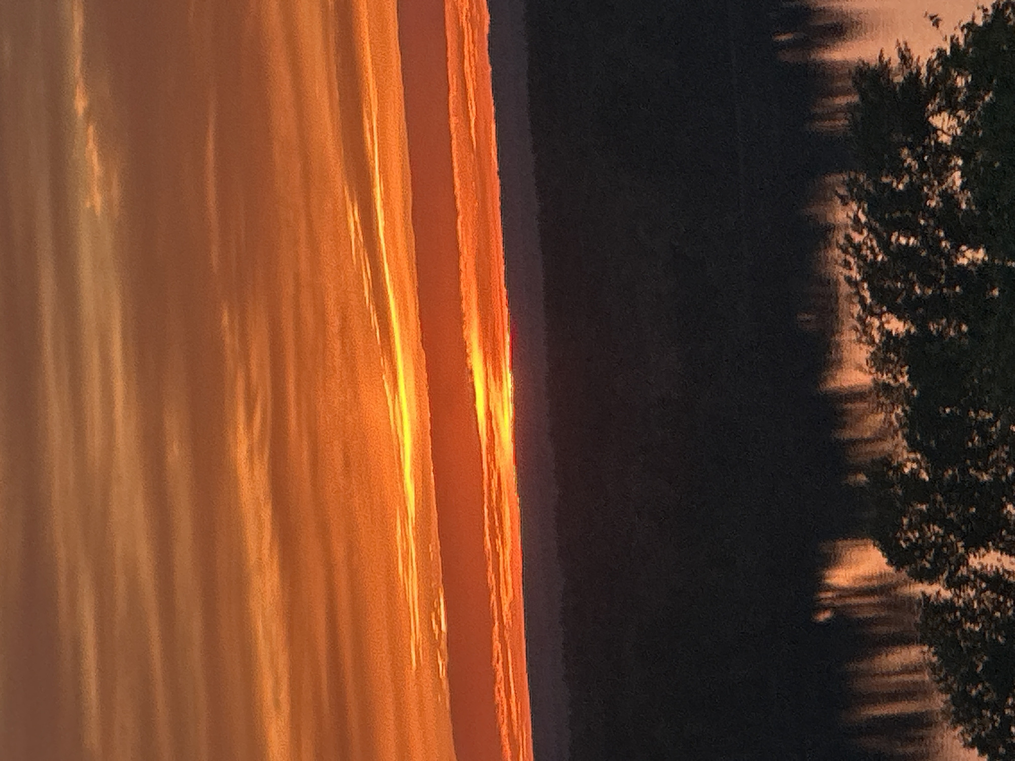 The image depicts a body of water at sunset, possibly the Kama River in Chaikovsky, Russia. The setting sun reflects on the water's surface, creating a bright orange and yellow glow. A tree line is visible on the right, its silhouette darkened against the water and sky. The sky is dark. The image appears to be taken from an elevated position.