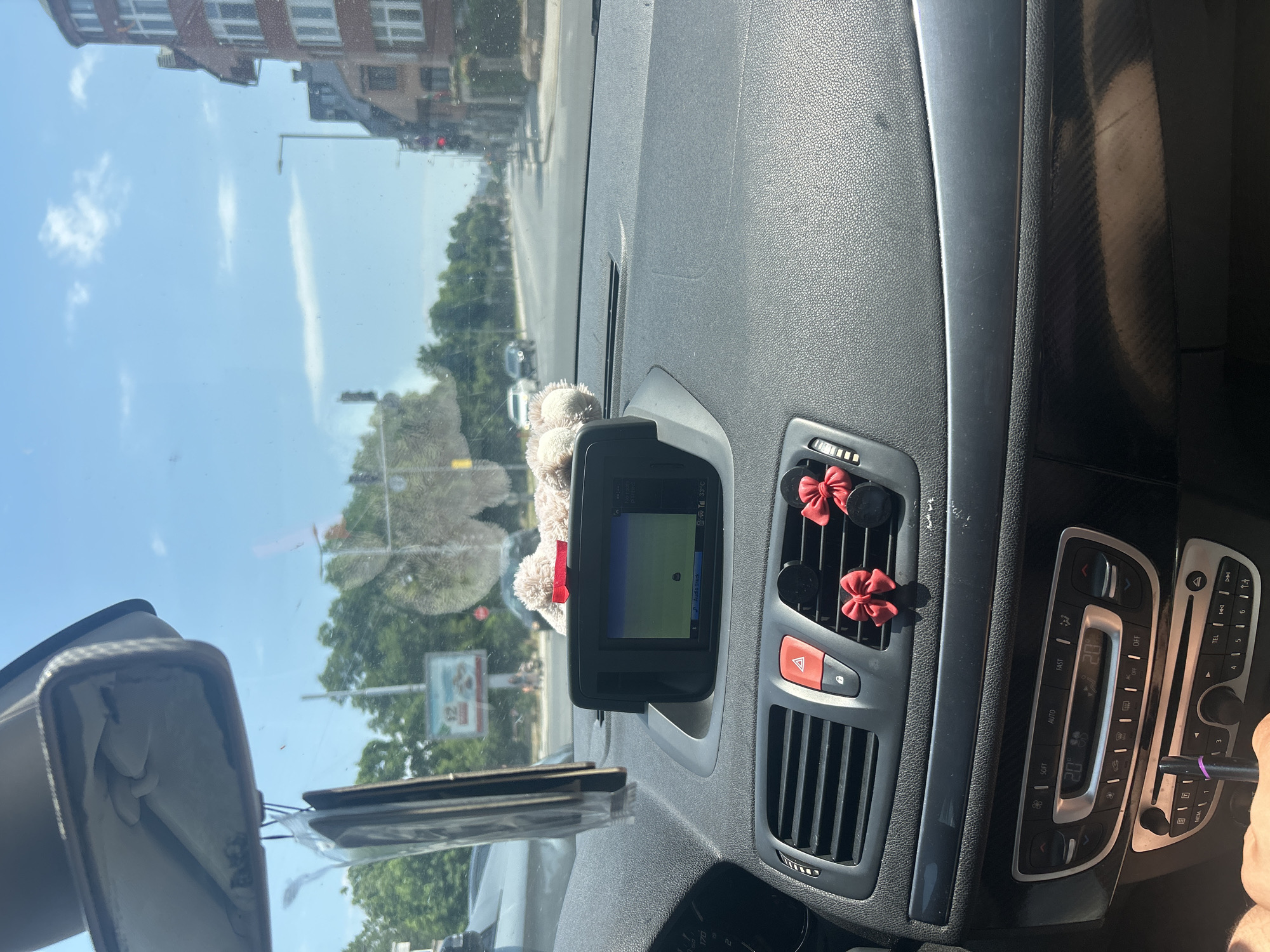 The image shows the interior of a car driving on a street in Plovdiv, Bulgaria. The viewpoint is from the passenger seat, looking forward. On the dashboard, a GPS screen displays a map, and two small plush teddy bears with red bows are attached to the air vents. The car's climate control system is visible below the GPS. Outside the car, a street lined with buildings and trees can be seen, with traffic lights and other cars on the road. The sky is blue with some clouds, indicating a sunny day.