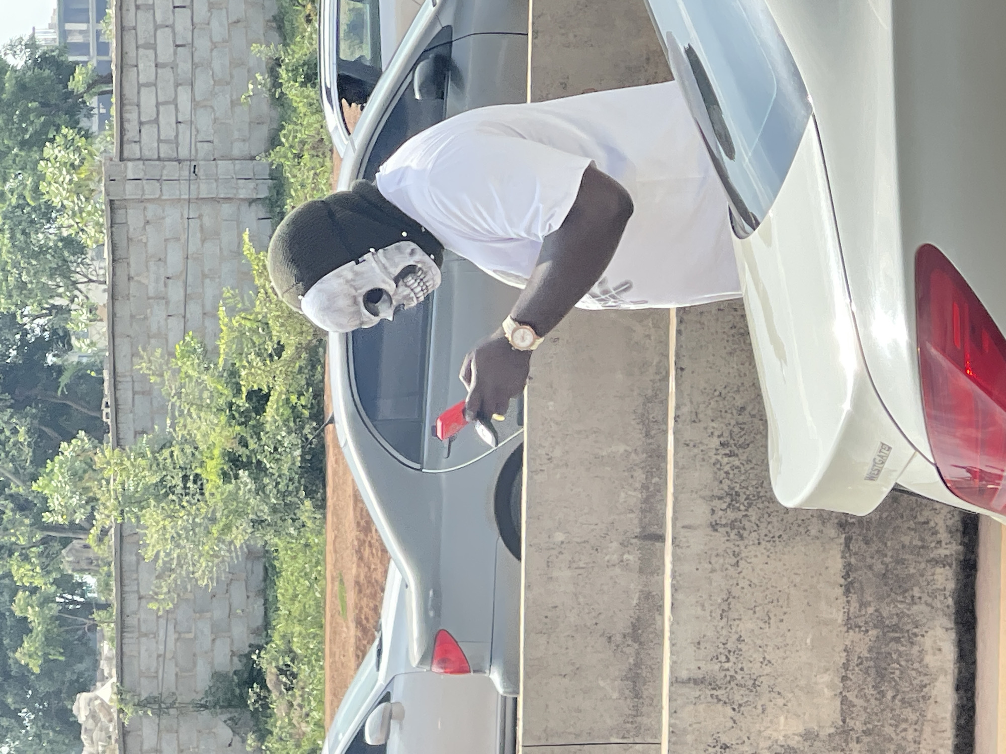 An individual wearing a skull mask and a dark head covering stands on a median strip beside a highway, holding a red mobile phone and a set of keys. The person is dressed in a white t-shirt and a gold wristwatch. Behind the individual, several vehicles are visible on the highway, including a gray sedan and a white sedan. The background features a concrete wall and lush green vegetation, characteristic of the landscape in Abuja, Nigeria. The lighting suggests it is daytime.