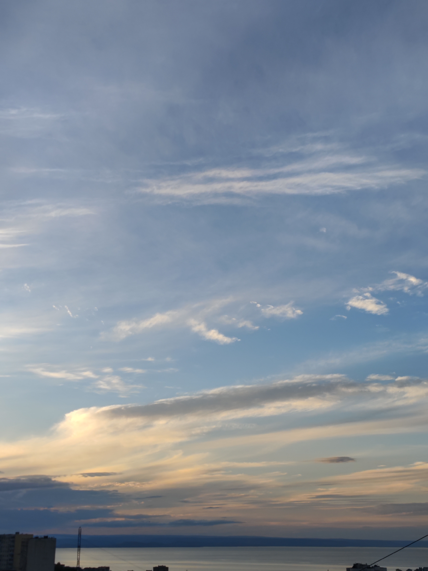 The image captures a wide view of the sky over Vladivostok, Russia, with a body of water and distant city structures visible in the lower portion. The sky is a prominent feature, characterized by a mix of light blue and grey hues, with scattered clouds. Wispy cirrus clouds streak across the upper half of the frame, interspersed with larger, more textured cumulus clouds in the lower half. The lower part of the image reveals a calm, expansive body of water, likely a bay or the sea, reflecting the muted colors of the sky. Along the horizon, the silhouettes of buildings and urban infrastructure are discernible. The lighting suggests it is either dawn or dusk, with the sun low on the horizon, casting soft, diffused light. No people or specific actions are depicted.