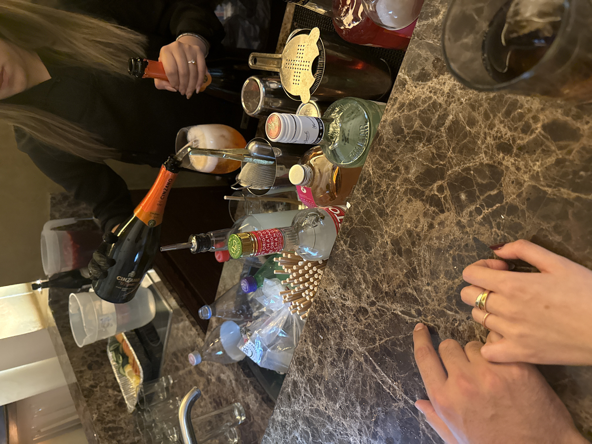 Here is a detailed description of the image:
**Scene:** The image appears to be taken at a bar or a domestic setting used as a bar, judging by the variety of alcoholic beverages, bar equipment, and the preparation of drinks. The countertop has a brown marble-like finish.
**People:** At least two people are present. One person, possibly a bartender or someone making drinks, is pouring from a bottle labeled "CINZANO". Another person, possibly a customer, is visible with their hands resting on the countertop.
**Subjects:** The main subjects are:
- The alcoholic beverages, including different bottles of liquor and wine.
- The bar equipment such as strainers and pour spouts.
- The hands of the people involved in the activity.
**Objects:** Numerous objects are scattered on the countertop, including:
- Bottles of alcohol (CINZANO, wine bottles, and other liquor bottles).
- Bar strainers.
- Pour spouts.
- A glass with a dark-colored drink and ice.
- Straws.
**Actions:** The primary action depicted is the pouring of liquid from a CINZANO bottle into a glass, presumably to mix a drink.
**Interactions:** The interaction focuses on making a drink with the bartender pouring from a bottle.
**Environment:** The environment has a bar-like setting with a countertop and the presence of various liquors and bar tools.
**Location Context:** There is no clear indication of Mexico City's location within the image.