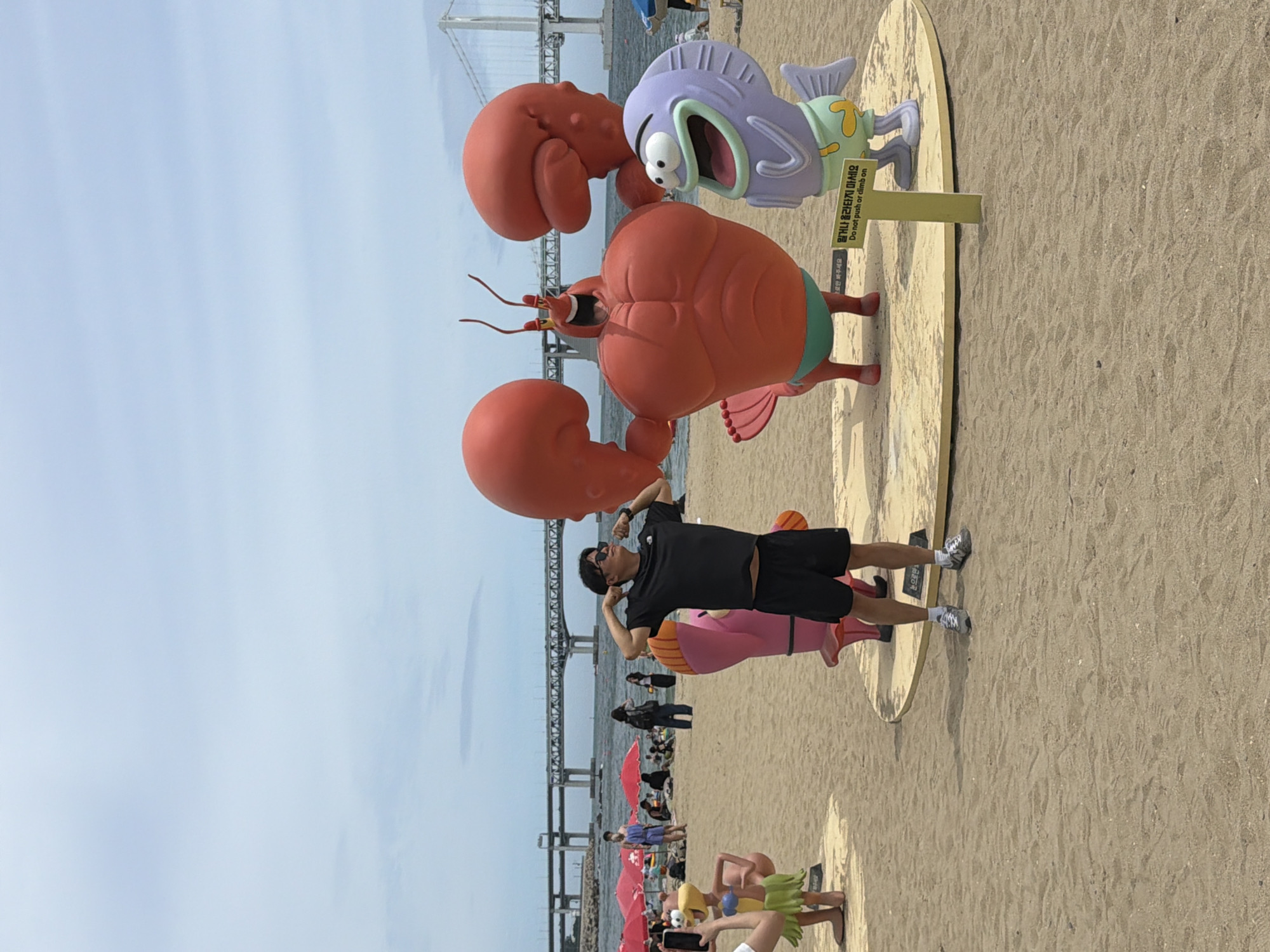 This photograph captures a scene on Haeundae Beach in Busan, South Korea. In the foreground, a man in a black t-shirt and black shorts is posing, flexing his biceps. He is wearing sunglasses and grey sneakers. Behind him are several large, colorful cartoon character statues. To the left is a pink figure, and to the right are two larger figures: a muscular red lobster with its arms raised and a light purple fish with wide eyes and an open mouth. The statues are standing on a raised, circular platform. A yellow sign with Korean and English text is posted in front of the fish statue, reading "Do not push or climb on." The beach is sandy, and in the background, a large bridge spans the water, with other beachgoers and red umbrellas visible further down the beach. The sky is overcast.
