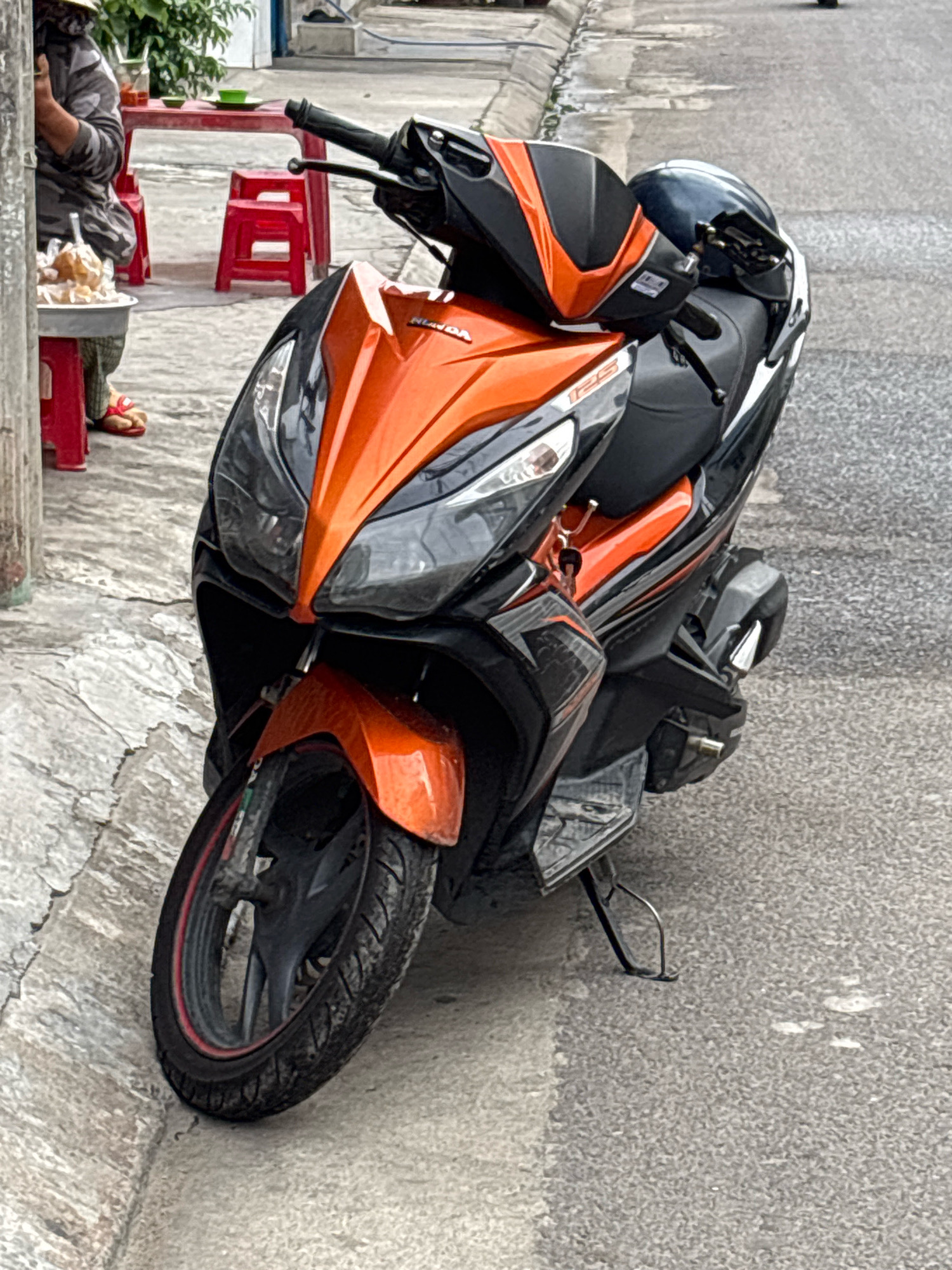 An orange and black Honda scooter is parked on the right side of a paved street, near the curb. A black helmet rests on the seat of the scooter. In the background, a person wearing a gray jacket and a hat sits at a small red table with red stools. A tray of food is on a small red stool in front of the person. The setting is a street in Nha Trang, Vietnam.