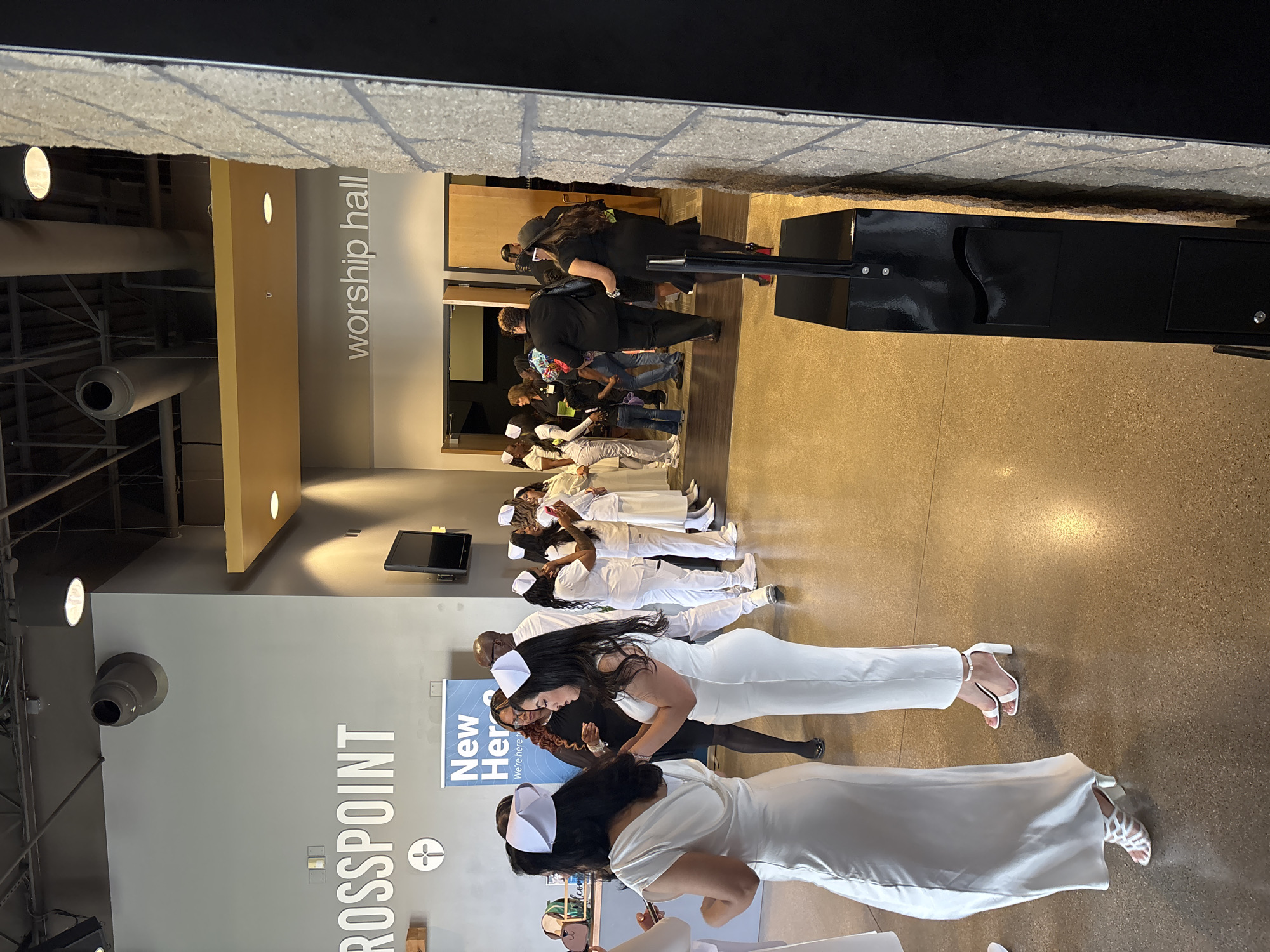 This media file captures an indoor gathering in Grand Prairie, United States of America, likely within a church or community center. The scene is dominated by a group of individuals, predominantly women, dressed in white attire, including dresses, pantsuits, and scrubs, many wearing white caps resembling traditional nurse caps. They are arranged in lines, suggesting an organized event or ceremony, possibly a graduation or induction for healthcare professionals, given their uniform appearance.
In the foreground, two women in white dresses and caps are visible, one adjusting her attire. Further back, more individuals in similar white outfits are lined up. In the background, other people are seen in casual, darker clothing, observing the event or waiting.
The setting is a modern indoor space with light-colored walls (grey and yellow/beige) and a polished concrete or light brown floor. The ceiling features exposed industrial-style structures and circular light fixtures. Visible on the wall is a large sign with the text "CROSSPOINT" and a cross symbol, indicating a church. Another banner features the text "New Heroes" and "We're here to". In the background, a sign reads "worship hall," confirming the nature of the venue. A black podium or stand is also present. The lighting is bright and artificial, suggesting an indoor event at an indeterminate time of day. The atmosphere appears formal and celebratory.