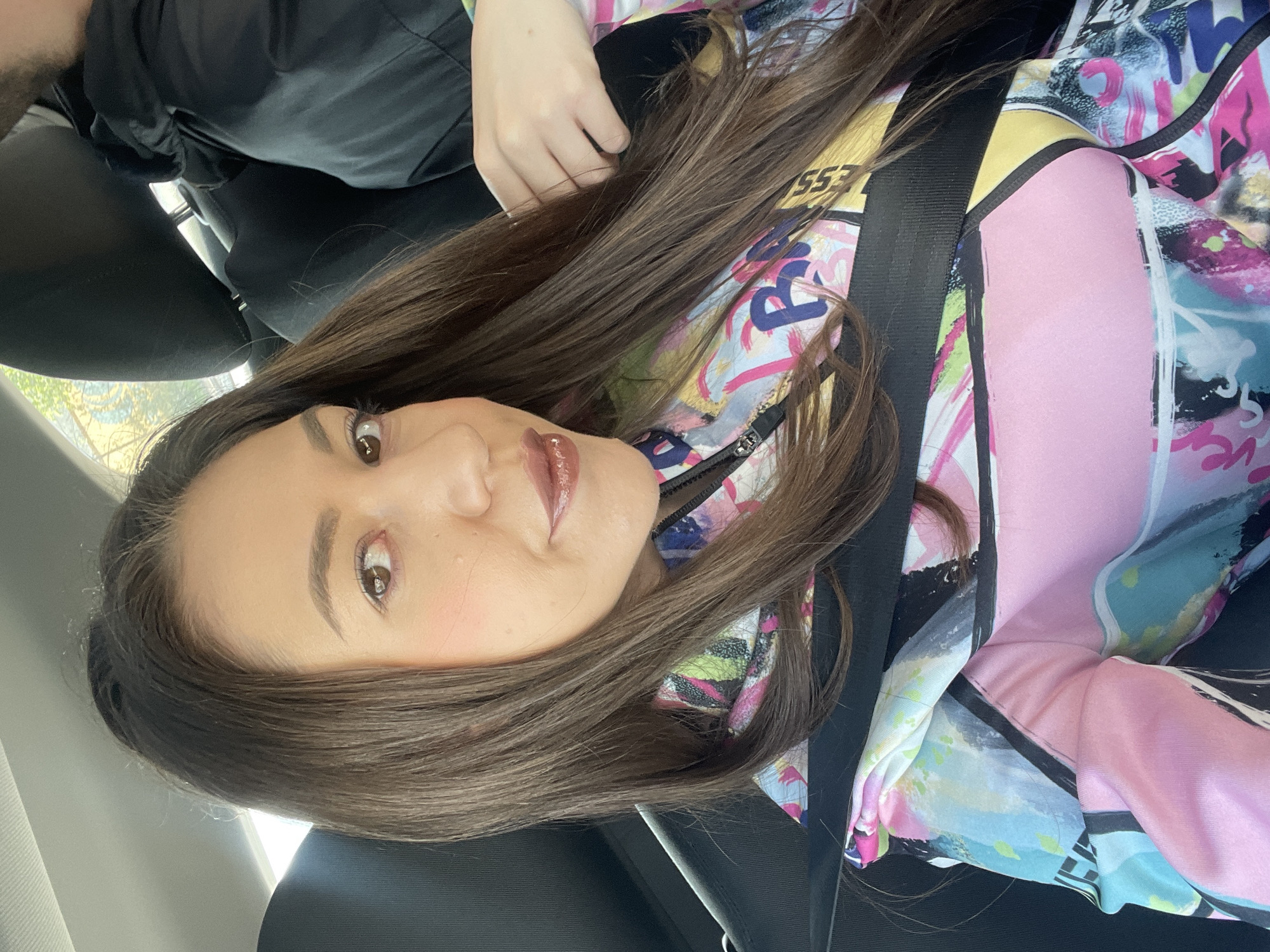 A young woman is seated inside a car, viewed from a slightly elevated angle with the image rotated 90 degrees clockwise. She has long, straight, dark brown hair that frames her face and flows over her shoulders. Her face is turned towards the camera, showing brown eyes and a neutral expression with glossy, parted lips. She is wearing a brightly colored jacket or hoodie featuring a dynamic, abstract, graffiti-style pattern with bold strokes of pink, blue, yellow, black, and white. A black seatbelt is visible across her chest.
The background inside the car is dark, likely the seat and headrest. Through the car window on her left, natural daylight illuminates the scene, and green foliage is visible, suggesting it is daytime. A partial view of another person or a reflection in a black garment is seen in the top left corner. The woman has noticeable makeup, including eyeshadow, blush, and defined eyebrows. Text is integrated into the jacket's design, with partially visible segments like "ESS" and "RES" on a yellow portion. The setting is within a car in Sofia, Bulgaria.