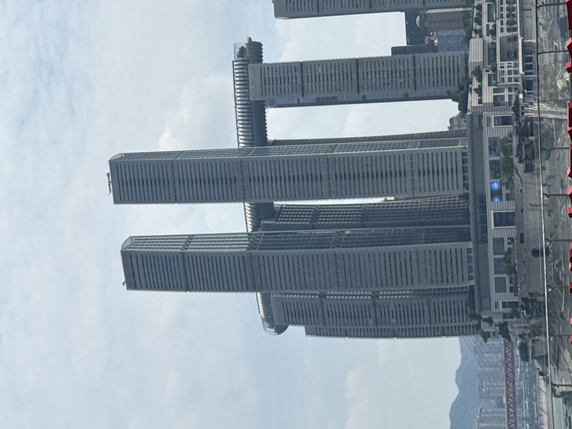 The image shows the Raffles City Chongqing complex in Nan'an District, China. The complex consists of eight skyscrapers, with two of the skyscrapers connected by a horizontal skybridge. The skyscrapers are tall and narrow, with a modern architectural style. The skybridge is a long, curved structure that spans between two of the buildings, featuring glass facades and what appears to be an observation deck. The buildings are made of glass and steel, with visible horizontal lines suggesting floors or windows. In the foreground, there are some red railings and a glimpse of a body of water. In the background, distant mountains and other buildings are visible, with a red bridge crossing a waterway. The sky is overcast with some light clouds. The scene appears to be a daytime, outdoor shot.