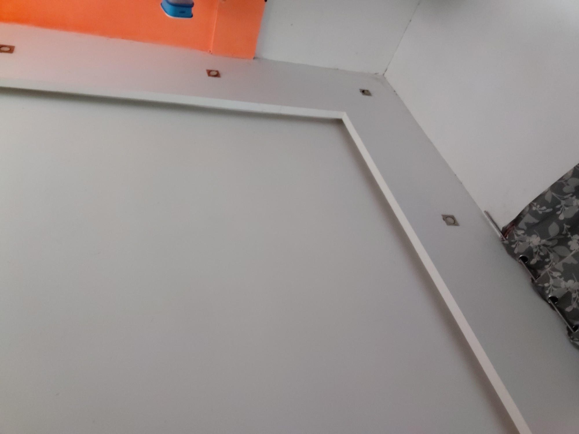 This image captures an interior view of a room, focusing on a white wall and ceiling. The main feature is a large, rectangular, recessed panel or framing on the white wall, occupying most of the lower and central part of the frame. Above this framed section, a white molding or soffit extends around the room's perimeter, leading up to the ceiling.
Embedded within this white molding are several small, dark, square or rectangular recessed light fixtures, appearing as spotlights. Three are clearly visible along the top edge of the white frame, and another two are visible along the right-hand molding. The upper part of the image shows a contrasting orange-colored ceiling or upper wall section. A light blue object, possibly part of a lighting fixture or utility box, is attached to this orange surface in the top-left corner.
In the top-right corner, a dark gray curtain with a white floral or leaf pattern is visible, hanging from rings. The room appears to be well-lit, likely by artificial light sources. No people or activities are visible. The overall aesthetic suggests a modern and minimalist design. The image is taken from a low angle, looking up towards the wall and ceiling. No visible text is present. The location context provided is Santa Ana, Venezuela.