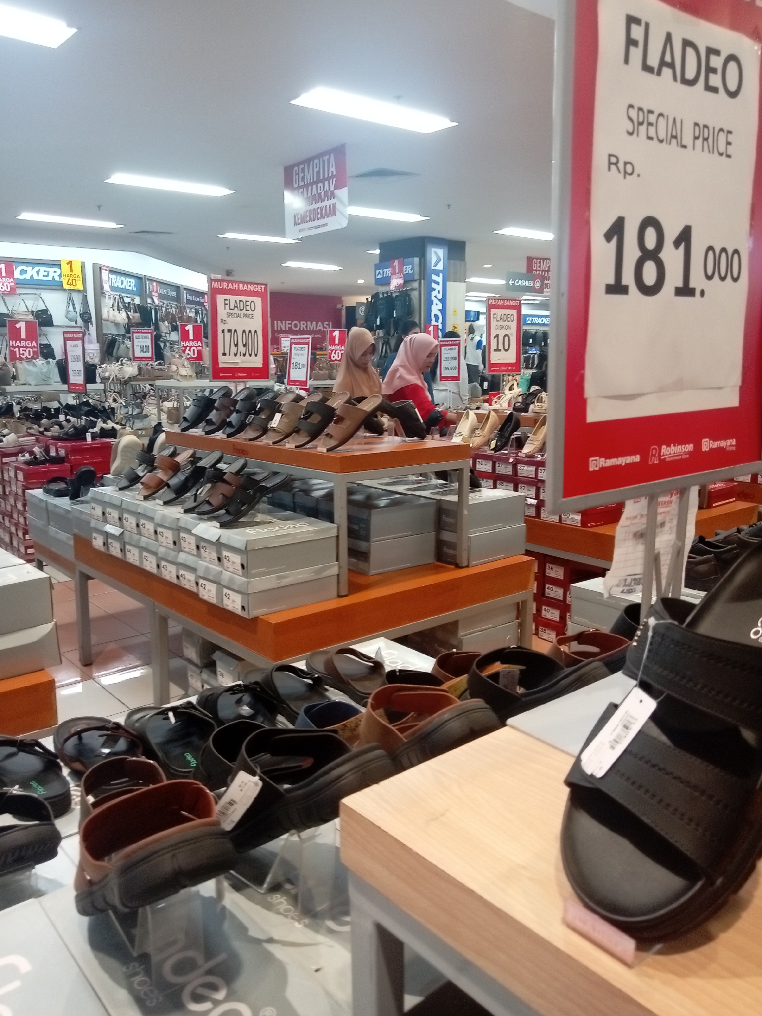 A sale is taking place at Robinson, a department store in Batam, Indonesia, as indicated by the store logos visible in the image. The scene focuses on the shoe section, with various display shelves and tables showcasing a range of footwear. A prominent red and white sign displays "FLADEO SPECIAL PRICE" with the price listed as Rp. 181,000. Another sign reads "FLADEO DISKON 10," suggesting a 10% discount. Several shoes, mostly sandals, are displayed on shelves and tables, with some stacked in boxes. Two women, one wearing a pink hijab and the other a beige hijab, are browsing the merchandise. The environment appears to be a well-lit indoor retail space with fluorescent lighting.