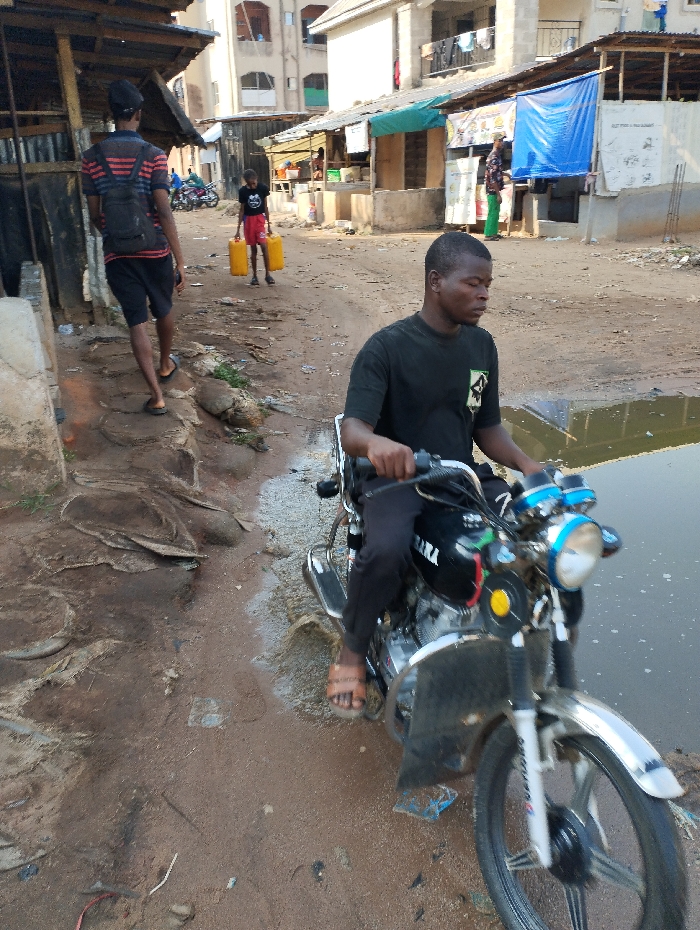 The scene depicts an unpaved street in Eziobodo, Nigeria, under daylight conditions. In the immediate foreground, an adult male operates a black motorcycle identified as "YAMAHA" brand, its front wheel partially submerged in a shallow water puddle on the dirt road. He wears a black t-shirt with a white chest graphic, dark trousers, and brown sandals, and is looking forward. The motorcycle features a chrome front fender, chrome engine parts, and a single headlight with blue trim.
To the mid-ground left, another adult male walks away from the camera, wearing a black baseball cap, a horizontally striped t-shirt (black and red), dark shorts, and black flip-flops, carrying a black backpack. Centered in the mid-ground, a young individual, likely a child or teenager, walks towards the camera, carrying one yellow plastic jerrycan in each hand.
The street surface is unpaved, consisting of packed dirt and mud, with multiple areas of standing water and exposed, hardened ground formations or roots along the left side. The background is populated by various structures: low-rise buildings constructed of concrete and wood with corrugated metal roofs, some featuring commercial signs and a prominent blue tarp draped over one section. Behind these, multi-story concrete residential buildings are visible, one with laundry hanging from an upper floor. Additional individuals and motorcycles are faintly discernible further down the street in the distance.