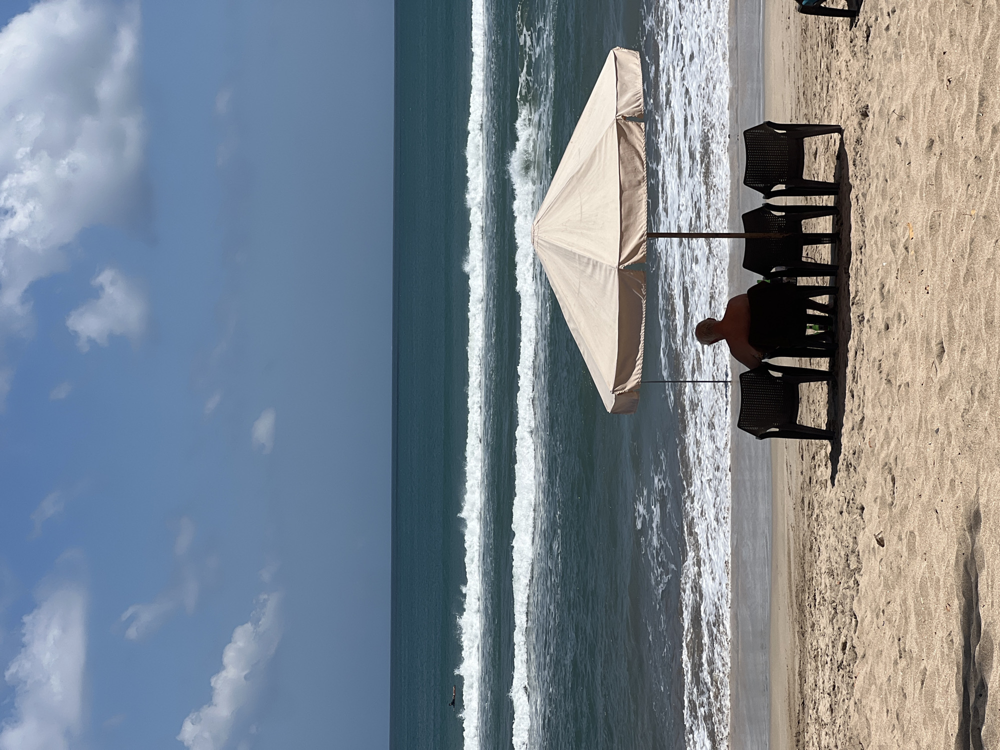 The image depicts a serene beach scene in Kuta, Indonesia, under a bright, cloudy sky. In the foreground, tan sand stretches across the beach, sparsely populated with a few dark plastic chairs arranged in a semi-circle. One chair is occupied by a man seen from behind, with white hair, facing the ocean. A large, light-colored beach umbrella provides shade over the chairs and the man.
Beyond the sand, the ocean stretches out, a deep teal color, with multiple sets of white-capped waves rolling towards the shore. The waves are consistent, suggesting moderate surf conditions. Far in the distance, a single surfer can be seen on a surfboard, paddling in the water. The horizon line is clear, separating the ocean from the pale blue sky, which is adorned with scattered white clouds. The overall atmosphere is one of tranquility and leisure.