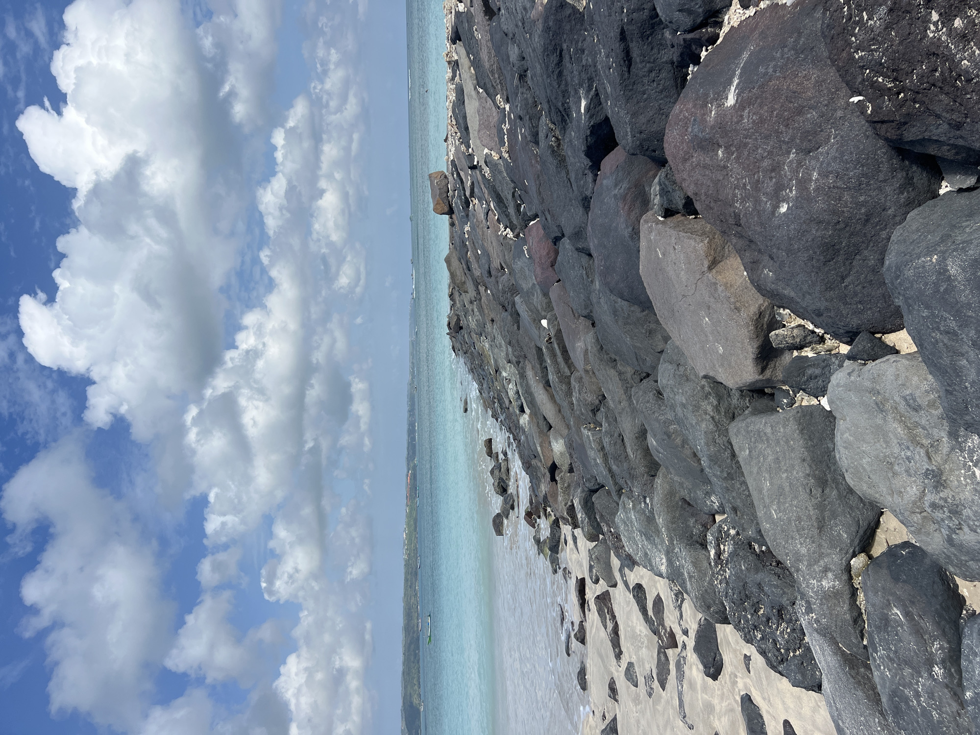 An eye-level, vertical, medium shot captures a rocky shoreline bordering a beach in Kuta, Indonesia. The foreground is dominated by large, dark, uneven volcanic rocks that form a breakwater extending from the right side of the frame towards the left. These rocks are interspersed with sand and occasional white debris, possibly coral fragments or shells. To the left of the breakwater, a sandy beach with scattered smaller rocks leads to the edge of the ocean. The water is a clear, turquoise hue, with gentle waves lapping at the shore. Further out, the ocean stretches to the horizon, appearing as a band of blue. In the distance, a faint coastline is visible, featuring green vegetation and scattered buildings. A few small boats are present on the water. The sky above is a vibrant blue, dotted with fluffy, white cumulus clouds. The scene depicts a natural coastal environment with man-made additions for erosion control.