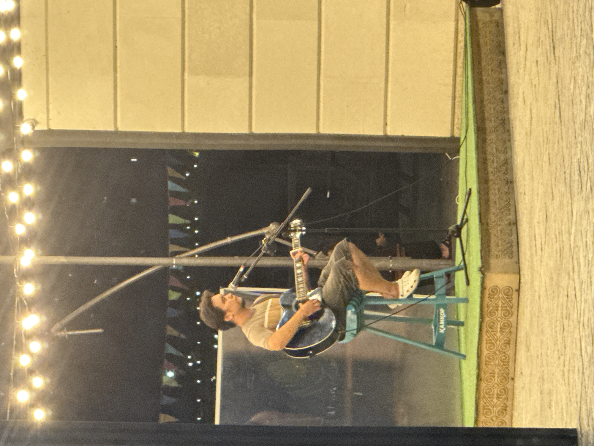 A man is playing an acoustic guitar and singing into a microphone while sitting on a teal stool on a raised stage. He is wearing a striped t-shirt and shorts. The stage is covered in green artificial grass and has a decorative wooden front. In the background, there are string lights and banners. The man appears to be performing outdoors at night. The location is likely the Embankment in Pavlodar, Kazakhstan, which is a popular public space that often hosts live performances.