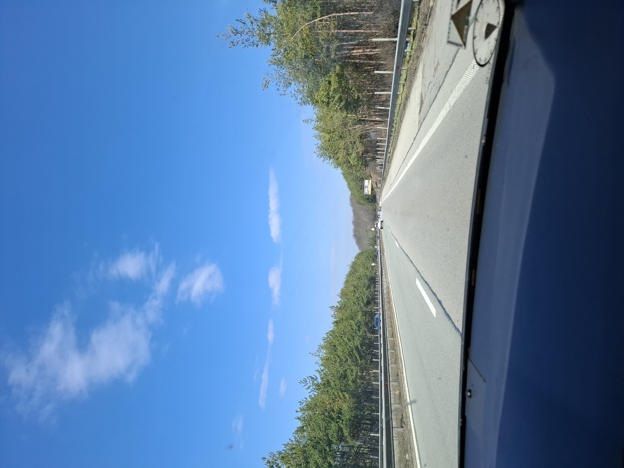 The image shows a view from inside a vehicle moving along a highway. The highway has two lanes and is bordered by trees and a metal guardrail. The sky is blue with some clouds. There are other vehicles visible in the distance on the highway. The view is from the passenger side of the vehicle. The location is inferred to be somewhere near Gorna Vasilitsa, Bulgaria, based on the prompt, although no details within the image directly confirm this. No people are clearly visible in the image.