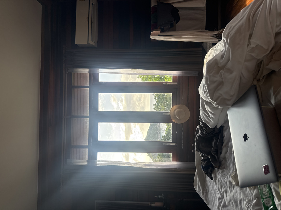 An indoor scene in Koh Tao, Thailand, features a room with dark wooden paneling on the walls and ceiling. In the immediate foreground, a disheveled bed with white and grey bedding is present. A silver Apple MacBook laptop, open and resting on a small stand, occupies the lower-left portion of the bed, accompanied by a green packet. A dark garment is crumpled on the bed's center.
A large, wooden-framed window dominates the midground, with bright sunlight streaming through its multiple vertical panes. A straw hat hangs centrally on the lower window frame. Translucent grey curtains are drawn to either side of the window. The exterior view reveals a tropical landscape with a prominent green, conical island rising from the sea under a partly cloudy sky. Lush green trees are visible closer to the window.
To the right, a white Mitsubishi air conditioning unit is mounted high on the wall. Below it, a clothes drying rack holds various garments, including white towels and dark clothing items. A partial view of a dark furnishing and a small table with bottles is visible on the far left. The scene is illuminated by strong natural light originating from the window.
