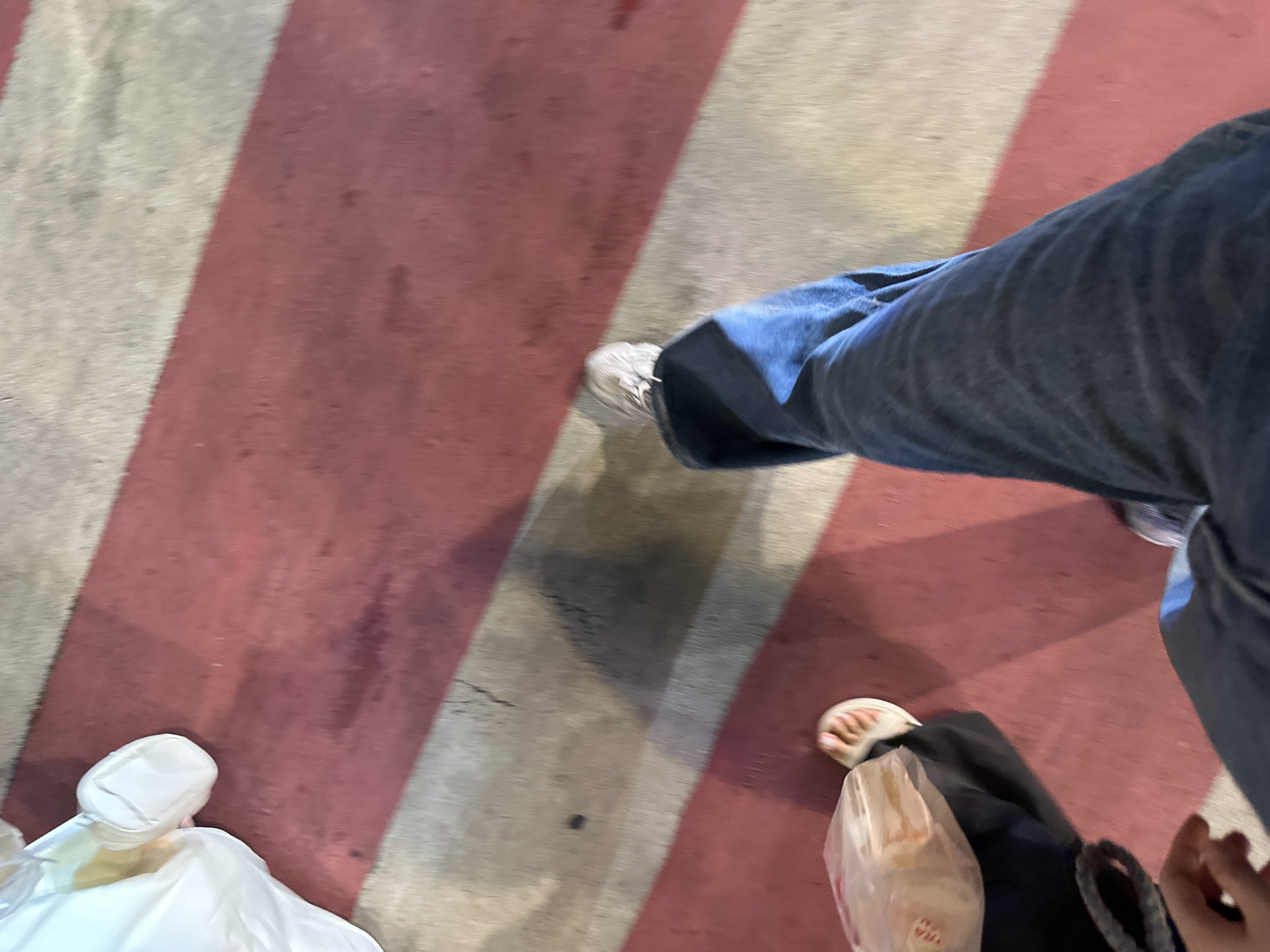 The image shows a pair of legs walking on a striped surface, which appears to be a pedestrian crossing. The stripes are red and white, and the ground is wet, suggesting it may have recently rained or the area is being cleaned. The person on the right is wearing blue jeans and white sneakers. The person on the left is wearing dark pants and a white sandal, carrying a plastic bag. The image is taken from a high angle, looking down at the legs and the ground. The lighting suggests it is daytime, possibly evening, given the shadows. The setting appears to be an urban environment in Thủ Đức, Vietnam. There is no visible text in the image. The overall impression is of people walking outdoors, engaged in everyday activity.