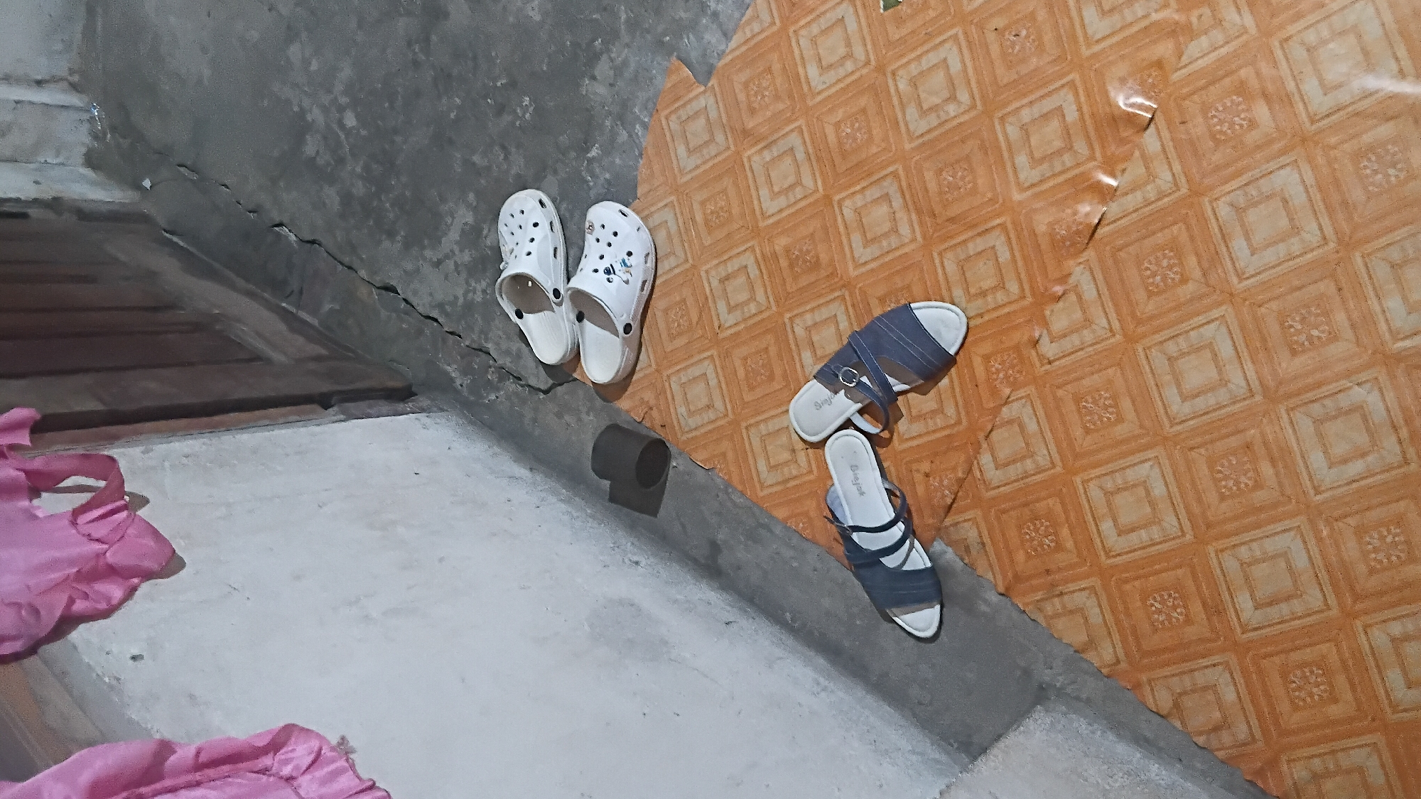 The image depicts a low-angle shot of a floor area with several pairs of footwear. In the foreground, on an orange-patterned mat, are two pairs of open-toed sandals. The sandals on the left are dark blue with white trim, and the pair on the right are also dark blue with white trim and a metallic buckle detail. Beyond the mat, on a concrete floor, are two pairs of white slip-on clogs. The clogs in the foreground have cartoonish designs, while the pair further back is plain white. To the left of the clogs, a dark gray cylindrical object is visible against a textured, light-colored wall. A folded piece of bright pink fabric hangs on the wall in the upper left corner of the image. The environment appears to be an indoor space, possibly a hallway or entryway, with a wooden door frame visible in the background and a crack in the concrete floor. The scene is well-lit, with no individuals present.