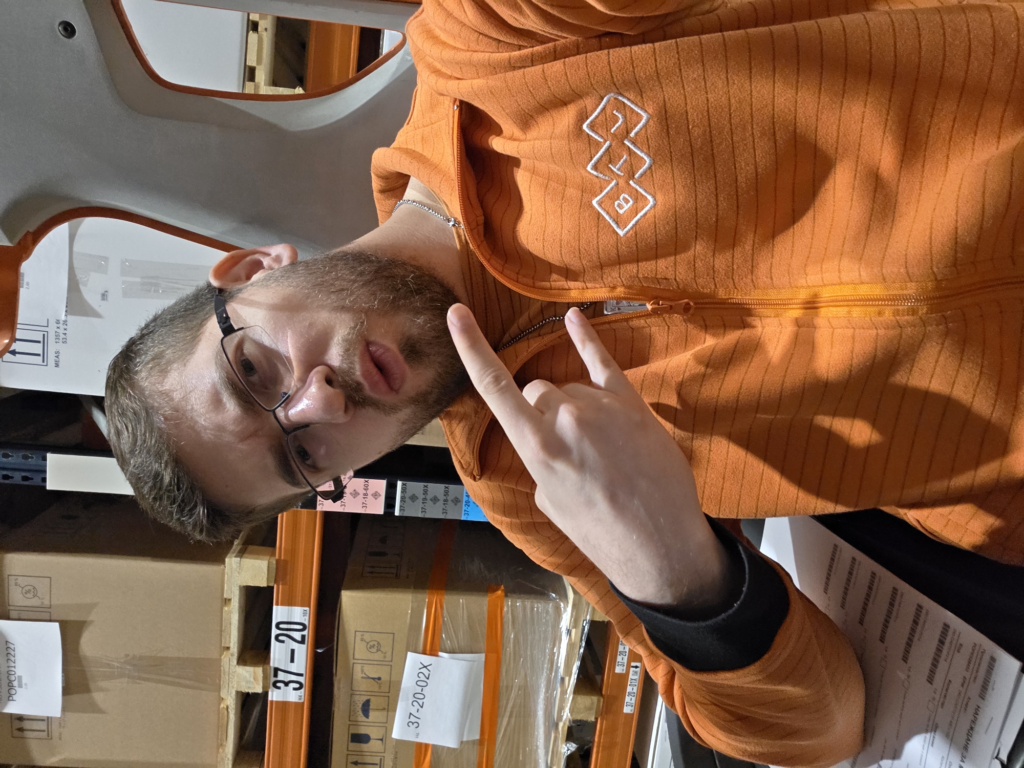 A man with glasses and a beard is pictured indoors, possibly in a warehouse environment, based on the visible shelving and stacked boxes. He is wearing an orange zip-up hoodie with a white logo. He is making a hand gesture with his index and pinky fingers extended. The boxes near him have labels with numbers such as "37-20" and "37-20-02X", as well as the phrase "НАРЕЖДАНЕ ЗА ГР," which could be Bulgarian. A sheet of paper lies next to the man, filled with what looks like barcodes and other printed information. Given the phrase "НАРЕЖДАНЕ ЗА ГР", which is in Bulgarian, the location may be Plovdiv, Bulgaria.