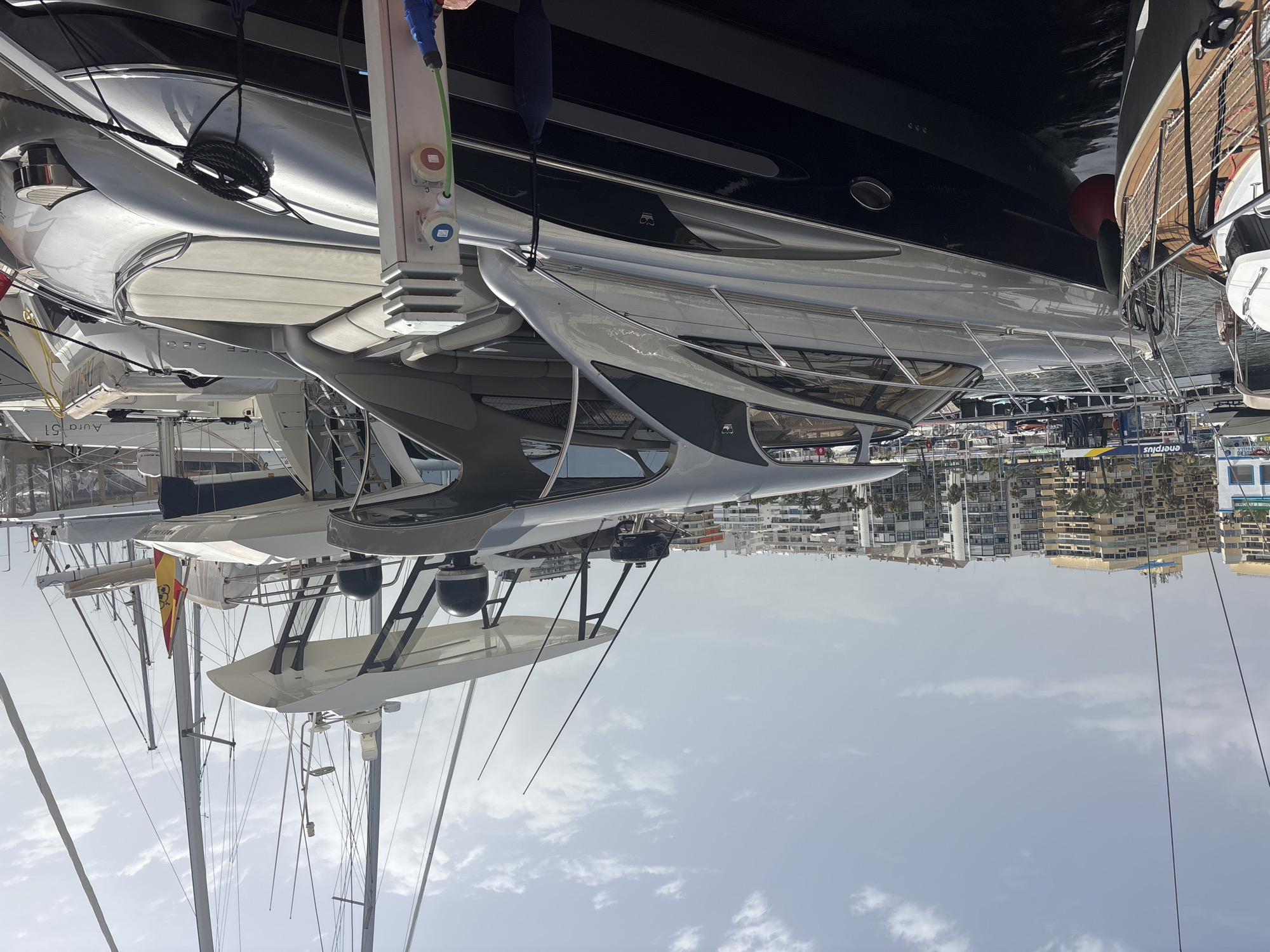 A high-angle, wide shot shows a luxury motor yacht docked in a marina in Marbella, Spain. The yacht, primarily silver and dark blue, is prominent in the foreground, with its sleek design and open seating area visible. It is moored alongside another, smaller white boat. In the background, several large sailboats with tall masts are docked, some with Spanish flags displayed. Behind the boats, a dense cityscape of apartment buildings and hotels lines the waterfront, with palm trees interspersed. The sky is partly cloudy, suggesting a warm, sunny day. Electrical connections are visible on a post near the yacht, with a green cable plugged in. There are no people clearly visible in the image.