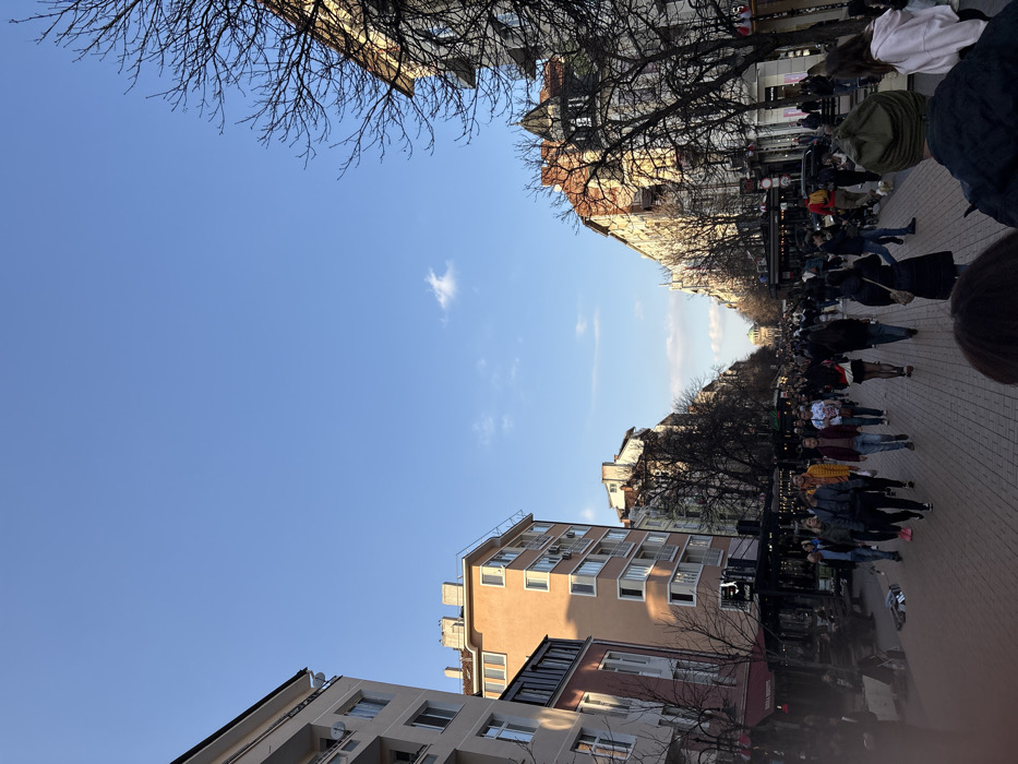 The image depicts a bustling pedestrian thoroughfare in Sofia, Bulgaria, identified as Vitosha Boulevard, oriented looking north towards the Largo complex. The scene is illuminated by a clear blue sky with sparse white clouds, indicating late afternoon or early evening sunlight, which highlights the upper facades of buildings on the right side. The street is paved with interlocking bricks and is densely populated with individuals of various ages dressed in casual attire, predominantly walking in both directions. Multi-story buildings with commercial establishments on their ground floors line both sides of the street. Buildings on the left feature lighter, modern facades, while those on the right display older, more ornate architecture. Deciduous trees, bare of leaves, punctuate the streetscape. In the far distance, the distinct dome of the National Assembly's former Party House building is visible. People are observed walking, congregating, and interacting casually. Outdoor seating for cafes is visible on the left.