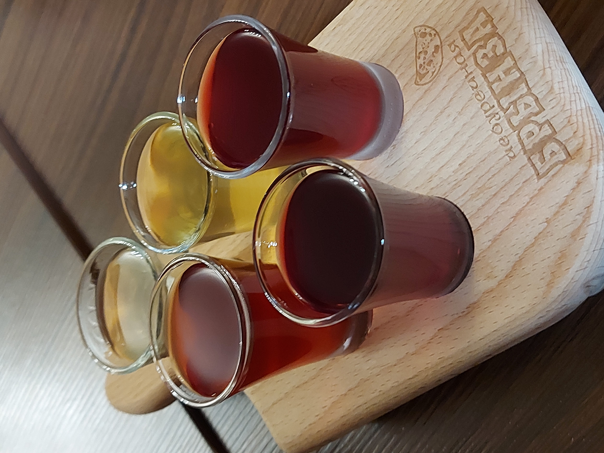 The image shows a wooden cutting board with six shot glasses filled with liquid. Four of the shot glasses are filled with a red liquid and two shot glasses contain a clear yellow liquid. The board has a logo imprinted on it, as well as the words "Десертная булочная." The location is Saint Petersburg, Russia.