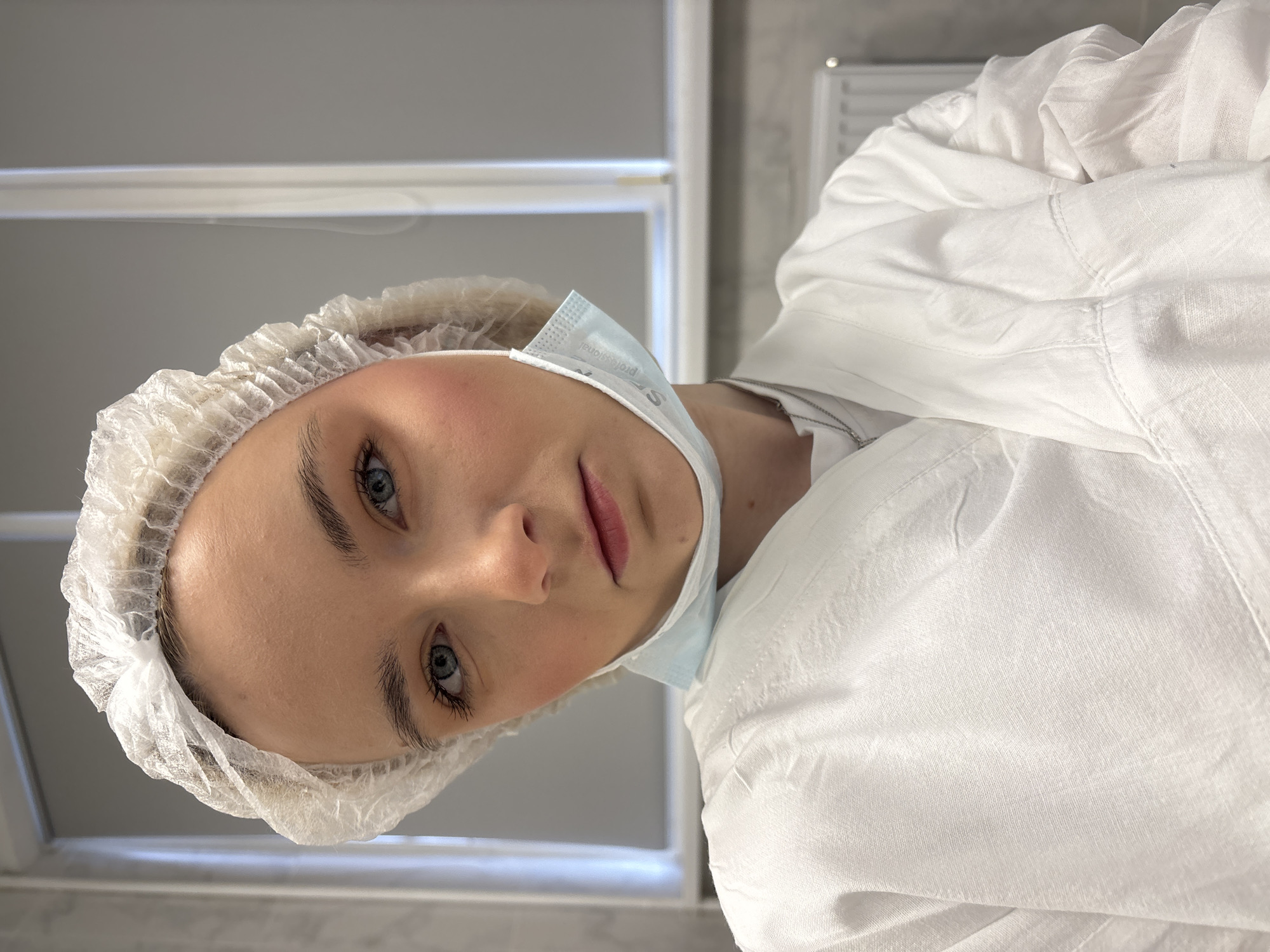 A young, fair-skinned woman with blue eyes is wearing a white bouffant cap and a light blue surgical mask pulled down below her chin. She is also wearing a white garment that resembles a lab coat or sterile gown. The background appears to be an indoor setting, possibly a medical or laboratory environment, with white or light-colored walls and what appears to be a fluorescent light fixture. The image appears to be rotated or taken from a low angle looking up at the woman. The location cannot be determined from the image.