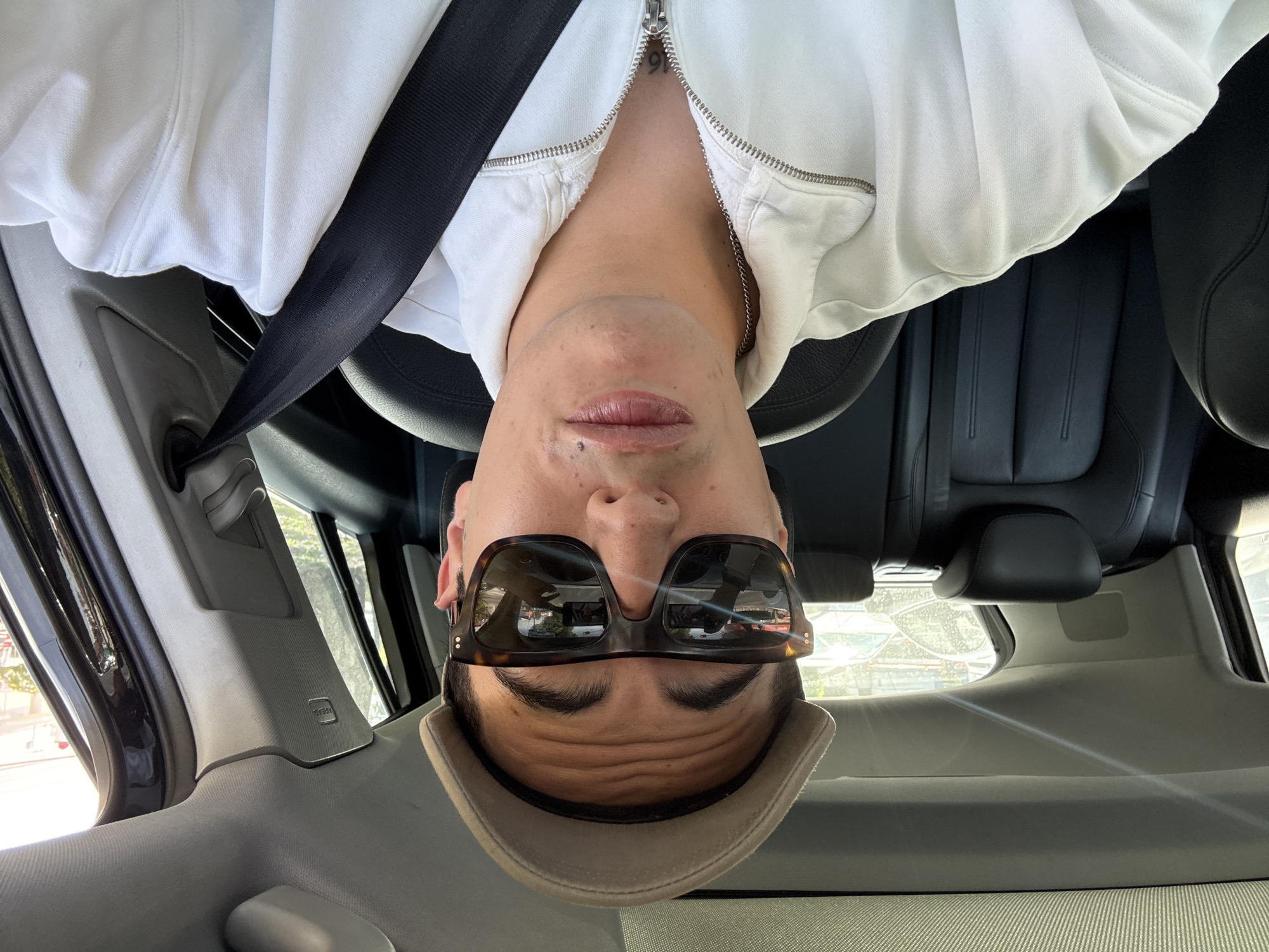 A person is positioned inside a vehicle, possibly a car, wearing a white zipped jacket and a hat. They are also wearing sunglasses and a seatbelt is visible. The interior of the car features dark leather seats and a light-colored dashboard. Through the car window, hints of a city landscape are visible. However, there is no conclusive evidence within the image to pinpoint the location as Puebla, Mexico.