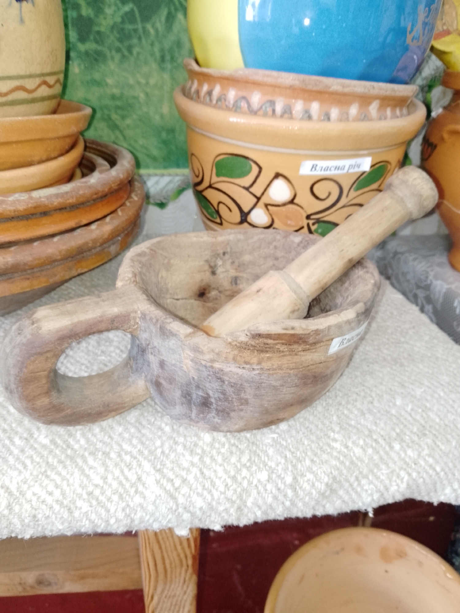 The image displays a collection of traditional Ukrainian crafts, likely from a market or shop in Ustyvytsia, Ukraine. The central focus is a rustic, weathered wooden mortar with a built-in handle, containing a matching wooden pestle. The wood exhibits visible grain and signs of age.
Arranged to the left of the mortar is a stack of circular, unglazed clay saucers or lids, showing a rough, earthy texture. In the background, several ceramic pots of varying sizes and colors are visible. A prominent light brown pot features painted green and dark brown decorative patterns. Attached to this pot is a small white label bearing the Cyrillic text "Власна річ," which translates to "Own thing" or "Personal item." A similar label is partially visible on the wooden mortar. Above the patterned pot, a vibrant blue glazed ceramic pot is partially seen, and to the far right, a section of an orange-brown pot is visible.
All items are displayed on a white, textured woven fabric, possibly a handmade tablecloth or mat, which rests on a light wooden surface. The background behind the pottery is a blurred green, hinting at a wall decoration or foliage. No people are present in the scene. The overall impression is one of traditional craftsmanship and local artistry, showcased in an indoor setting.