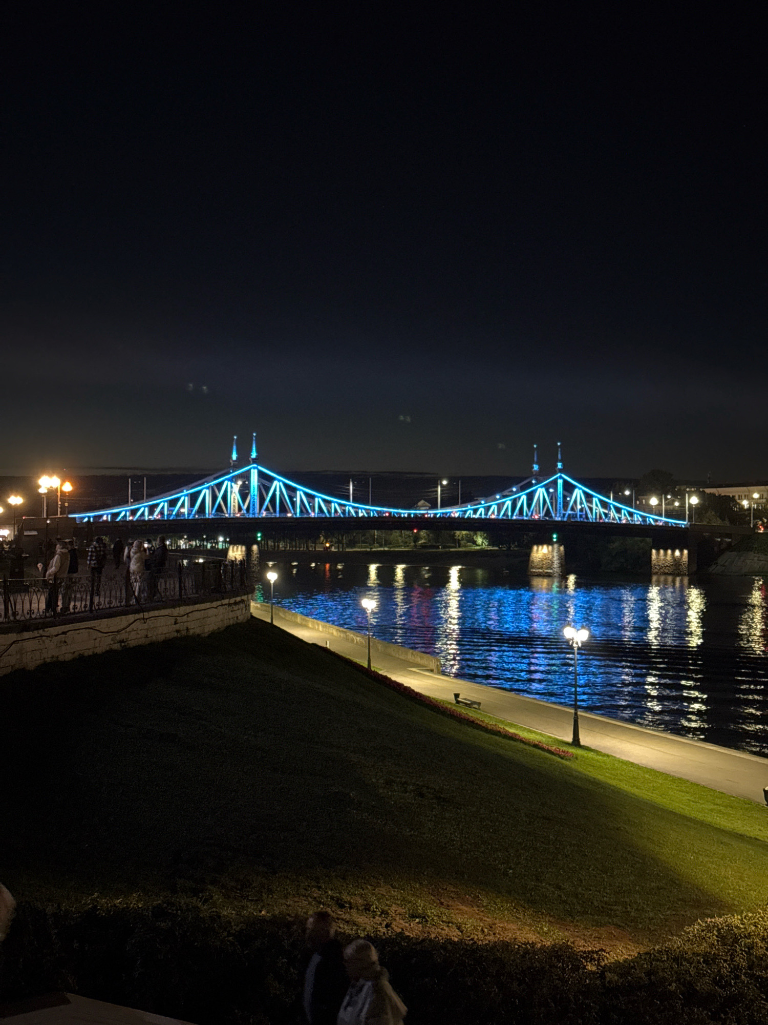 The image depicts a night scene of the Starovolzhsky Bridge in Tver, Russia, illuminated with vibrant blue lights. The bridge spans across the Volga River, and its elegant structure is clearly visible against the dark night sky. The water of the river reflects the bridge's lights, creating a shimmering effect.
On the left side of the image, a promenade or embankment is visible with several people walking along. Streetlights with warm yellow light dot the promenade, illuminating the pathway and the surrounding area. The embankment is covered in grass, and a stone retaining wall separates it from the river.
In the foreground, a grassy slope descends towards the river, with more people visible walking at the bottom of the slope. The overall atmosphere is serene and picturesque, highlighting the beauty of the city at night.
