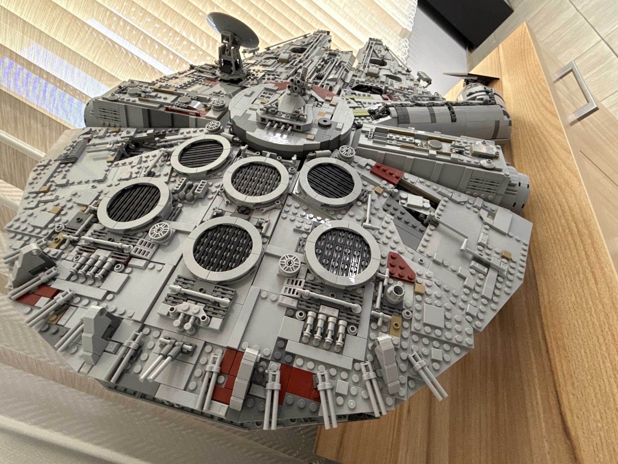 A high-angle, close-up shot depicts a detailed Lego model of the Millennium Falcon spaceship on a light-colored wooden surface. The model, primarily grey, exhibits intricate detailing across its surface, with circular features and an assortment of grey and red accents. A radar dish is situated on top. The backdrop consists of vertical blinds on the left and a wooden cabinet on the right, suggesting an indoor setting. The scene is not located in Krasnoyarsk, Russia.