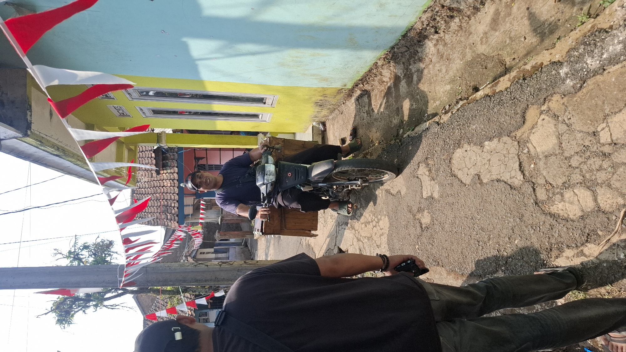 A man wearing a dark t-shirt and a baseball cap is seated on a motorcycle with a wooden box attached to the rear. He is smiling and looking at the camera. The motorcycle is parked on a paved road that appears to be in disrepair, with several potholes visible. The man is wearing dark pants and sandals.
To the left of the motorcycle, another person is visible from the back, wearing a dark t-shirt and a black baseball cap. This person is holding a black object in their right hand.
The scene is decorated with a string of red and white triangular flags, which are characteristic of Indonesian independence day celebrations. The flags are strung across the street, partially obscuring a view of houses in the background. The houses are painted in bright colors, with one house being yellow and another light blue. The street appears to be in a residential area.
The lighting suggests it is daytime, with sunlight casting shadows on the ground. The overall impression is a casual street scene in Indonesia, possibly during a festive occasion. The location is specified as Cilaku, Indonesia.