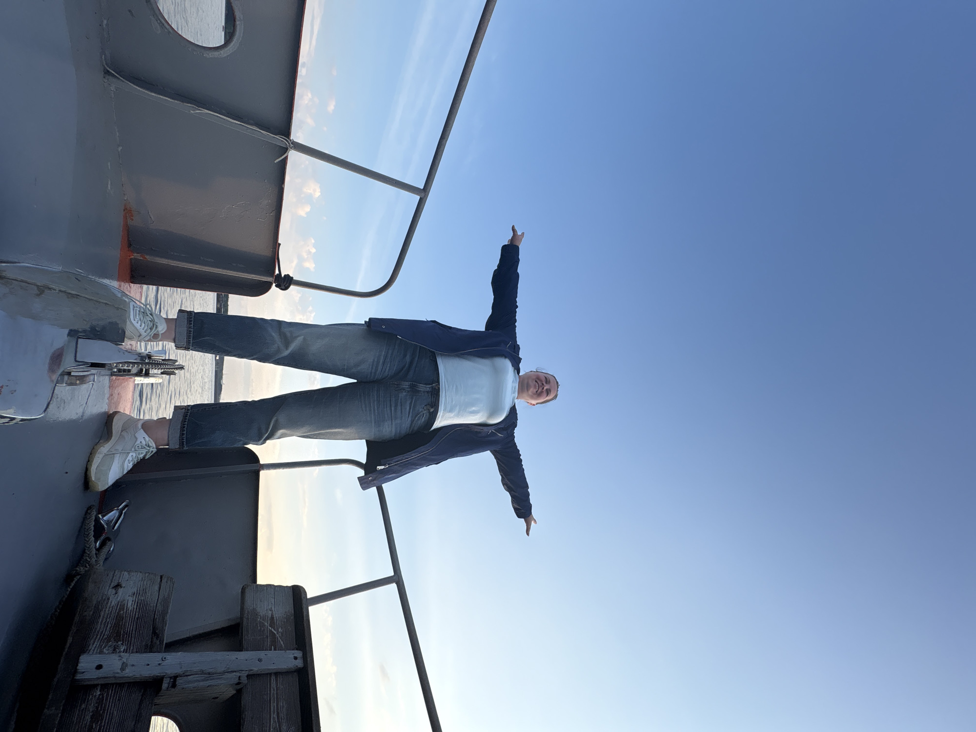 A person stands on the bow of a boat with arms outstretched towards a clear blue sky. The individual is wearing a light-colored t-shirt, a dark jacket, and jeans, with white sneakers. The boat appears to be in open water, with railings visible on either side and a glimpse of the water and distant shore. The lighting suggests it is either early morning or late afternoon. The image captures a moment of freedom and enjoyment on the water.