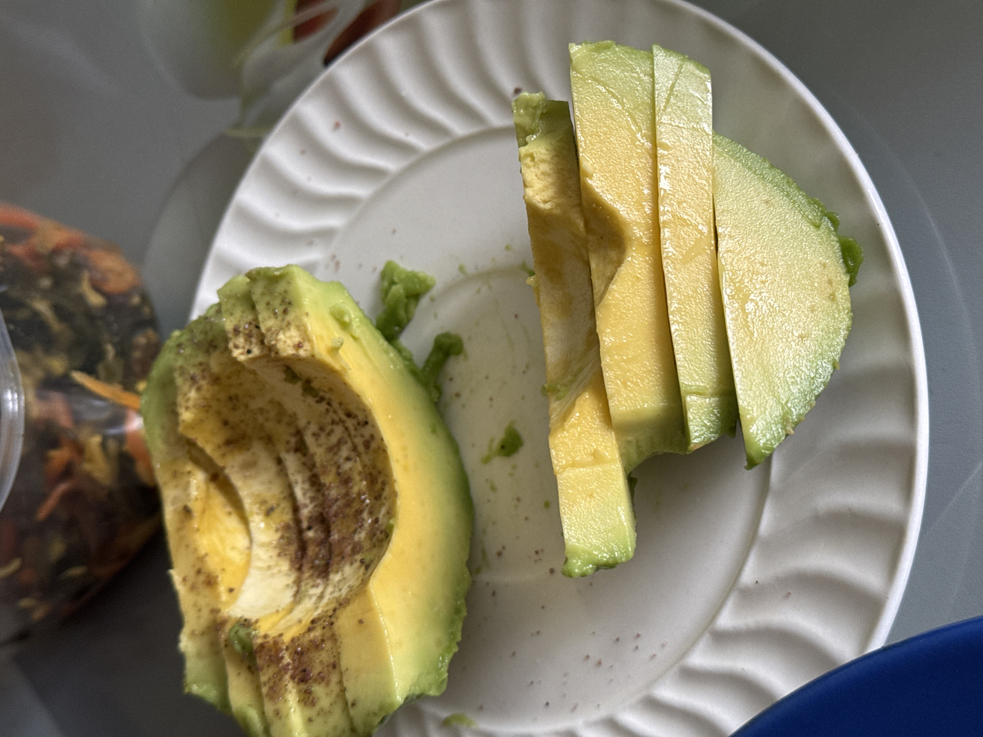 A close-up, overhead shot features a sliced avocado served on a white ceramic plate with a decorative wavy rim. The avocado is divided into two main parts: on the left, one half of an avocado, with its seed removed, lies flat on the plate, showing its pale yellow-green flesh dusted with a dark brown, speckled seasoning. On the right side of the plate, several long, thin slices of avocado are neatly arranged, standing upright, showcasing their vibrant green skin and creamy interior.
The white plate itself shows some scattered small brown specks, likely residue from the seasoning applied to the avocado. In the blurred background to the left, a translucent plastic container with a mix of dark and lighter-colored food items, possibly pickled vegetables or a salad, is partially visible. A hint of a blue object, likely another dish or bowl, is visible in the bottom right corner of the frame. The overall lighting suggests an indoor setting. No people or visible text are present in the image.