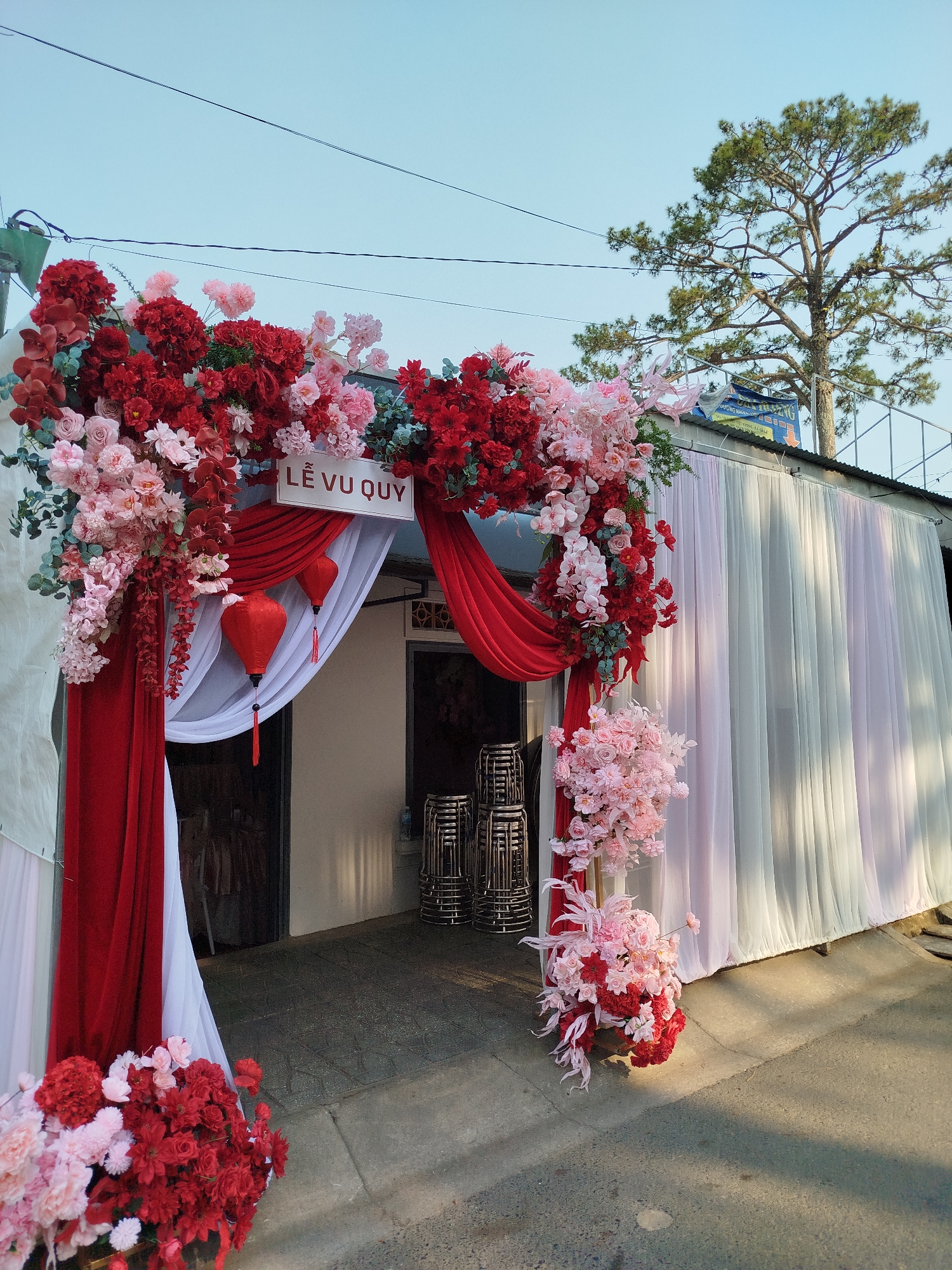 An outdoor wedding entrance in Da Lat, Vietnam, is presented under a clear daylight sky. A prominent archway, serving as the main entrance to a building, is elaborately decorated with dense arrangements of artificial flowers in shades of red, pink, and white, interspersed with green foliage. Red and white fabric drapes frame the archway, and a white sign reading "LỄ VU QUY" in black text is centered above the opening. Two red traditional Vietnamese lanterns with black tassels hang within the draped archway.
Behind the archway's drapes, a dark doorway is visible, leading into the building. Stacks of light-colored banquet chairs are positioned on the ground, both partially inside the entrance and to its immediate right. The building's exterior to the right of the archway is covered by vertical panels of sheer fabric in white and pale pink. Above this, a corrugated metal roof is visible, along with a partially obscured blue banner bearing white text. In the upper right background, a tall, mature pine tree stands against the light blue sky. Multiple thin black power lines traverse the sky horizontally. The foreground consists of paved concrete or asphalt ground, upon which distinct shadows are cast by direct sunlight. No individuals are present in the scene.